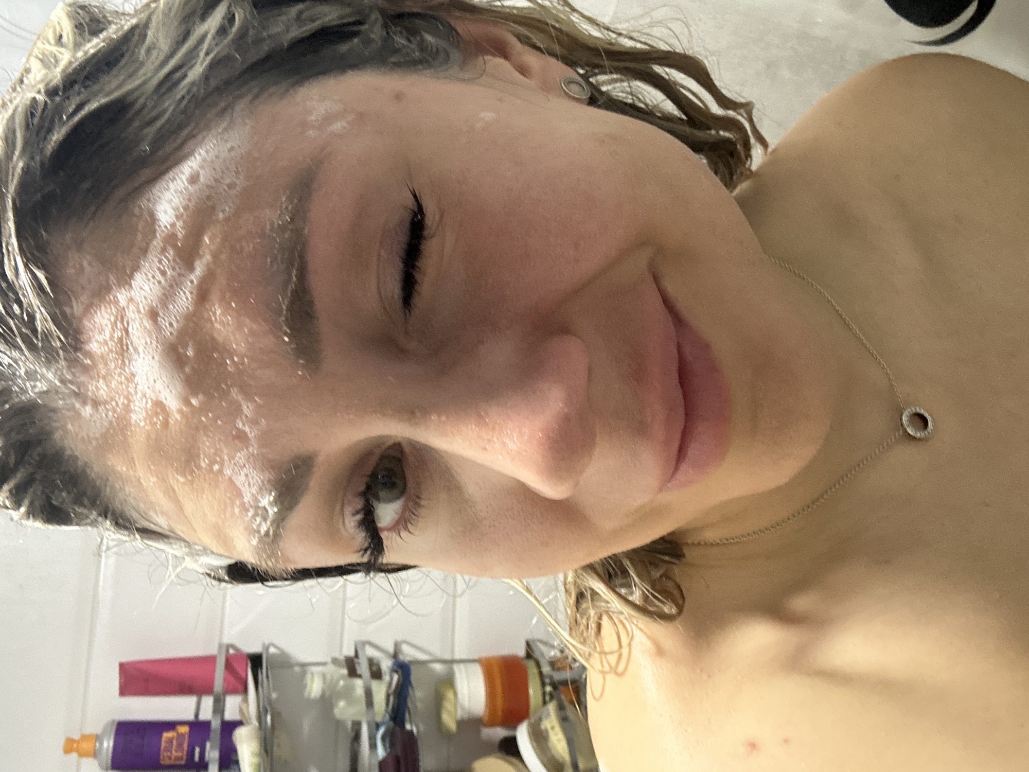 A close-up, eye-level, medium shot shows a woman in a shower, with her face covered in soap suds. She is winking at the camera. Her hair is wet and pulled back, and she is wearing a necklace. Behind her, shelves in the shower are stocked with various toiletries. The lighting is bright, and the background is a tiled wall. The scene is indoors, likely a bathroom. There is no information in the image to indicate its location in Novoaltaysk, Russia.