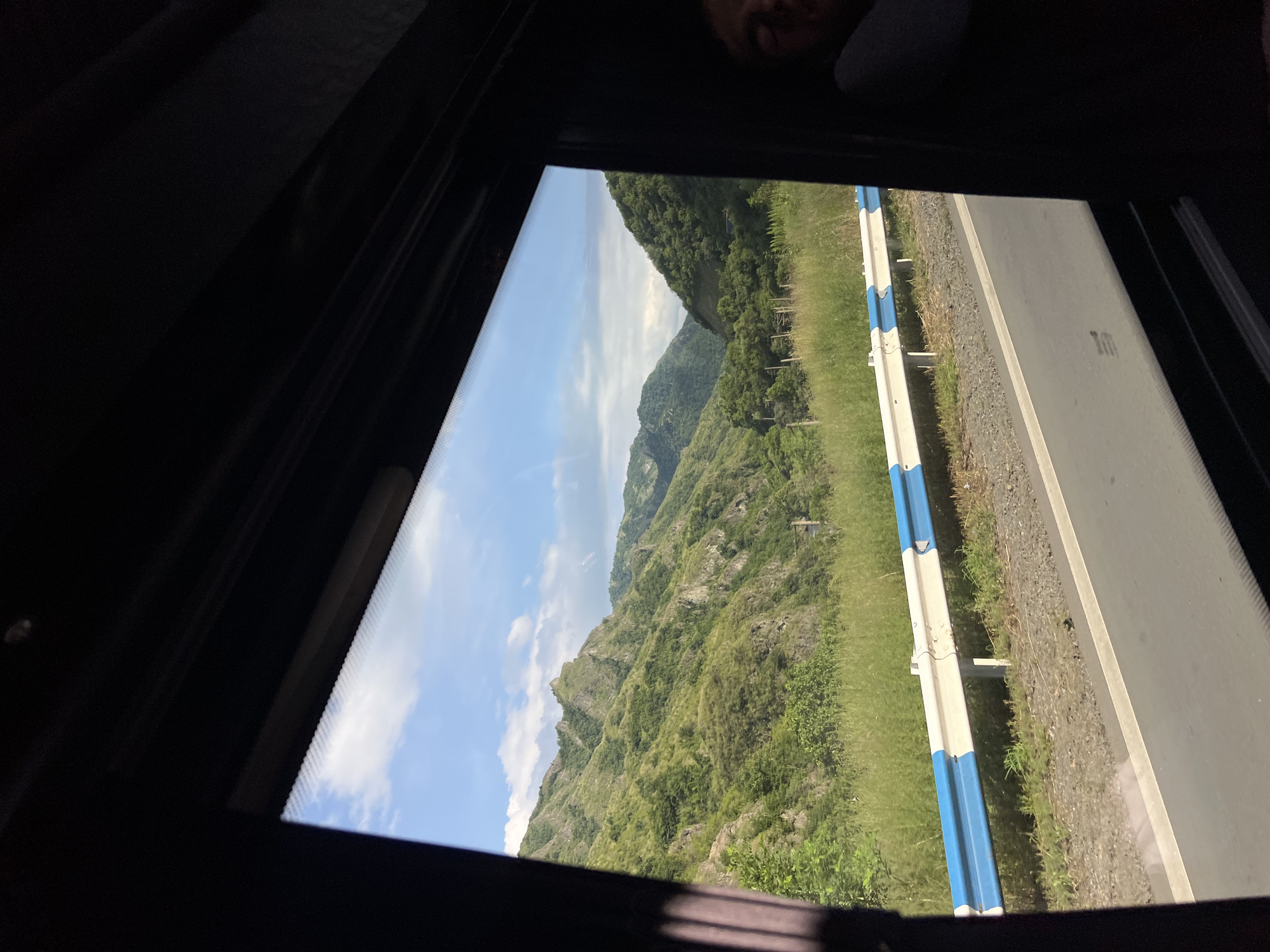 The image depicts a view from a window, likely from a vehicle, looking out at a mountainous landscape. The foreground is dominated by the dark interior of the vehicle, with a sliver of blue sky and white clouds visible at the top. The main subject of the image is a series of lush green mountains, with rugged peaks and dense vegetation. A paved road with a white and blue guardrail runs horizontally across the mid-ground. Tall, dry grass lines the side of the road. On the right side of the frame, a portion of a person's face, with eyes closed and head resting, is visible, suggesting they are asleep or resting. The overall scene conveys a sense of travel through a scenic, rural area. The location is identified as Odzun, Armenia.