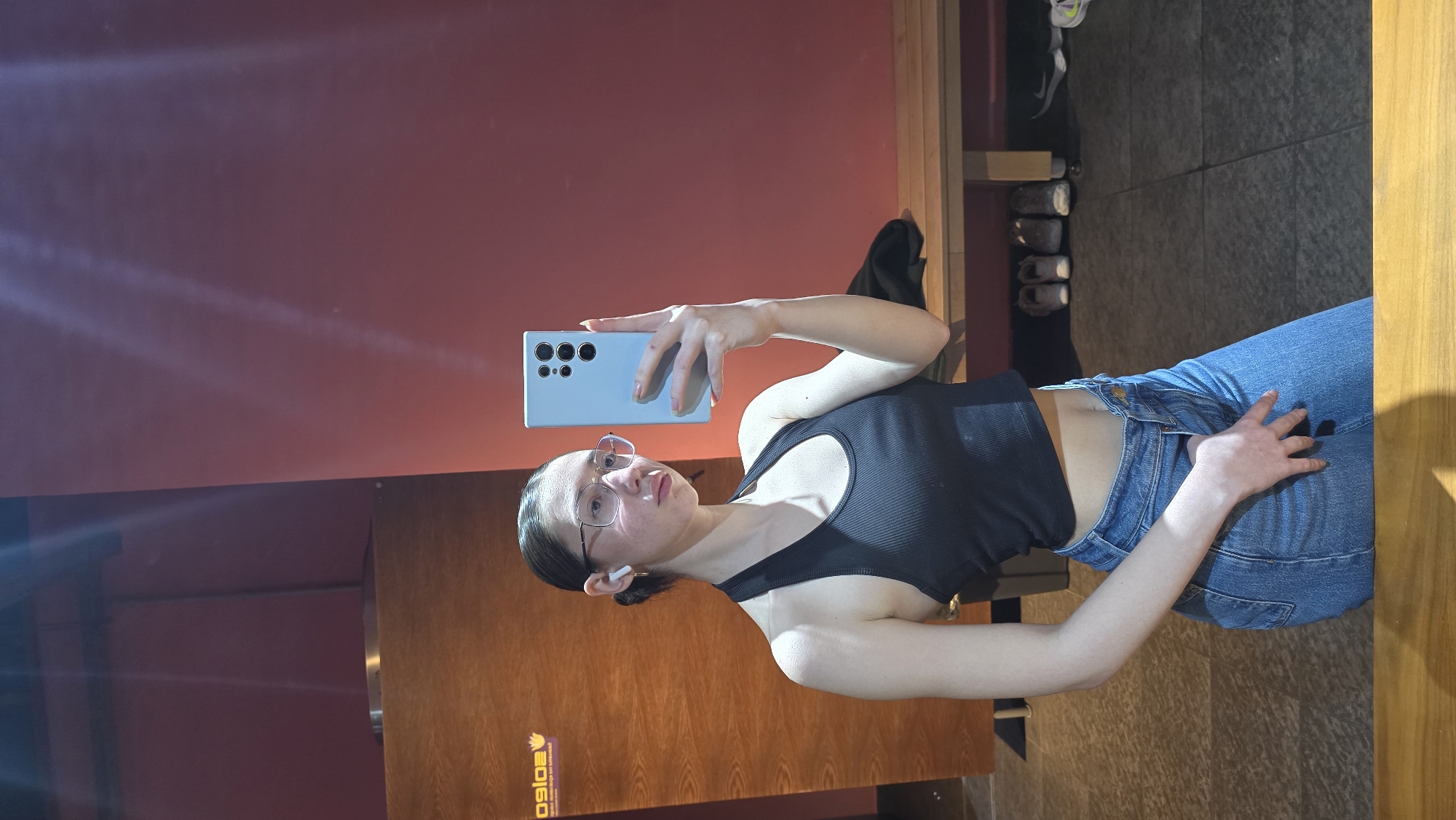 A woman stands in front of a mirror taking a selfie with a light blue smartphone. She is wearing jeans, a black tank top, and glasses. She has a white earbud in her left ear. The background includes a red wall and a wooden door with a logo on the lower left. The location is likely indoors, possibly a gym or studio in an unspecified location in the city of Stara Zagora, Bulgaria.