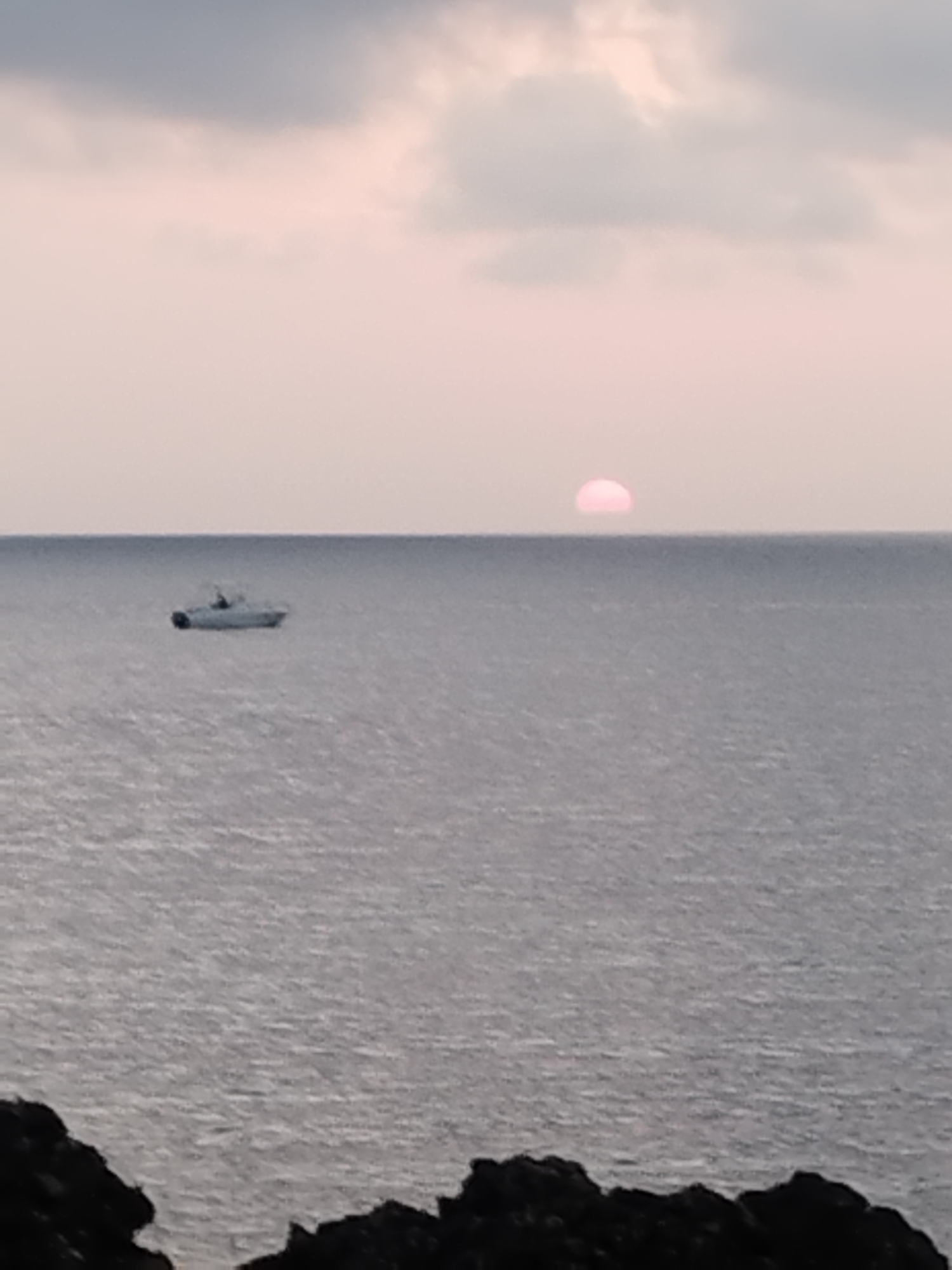 This image captures a serene seascape with a soft, pinkish sky, likely at sunrise or sunset. A crescent of the sun, also tinged with pink, is visible just above the horizon line. The vast expanse of the ocean stretches out, reflecting the muted light of the sky. A small, white boat is present in the mid-ground, floating calmly on the water. In the foreground, the dark, rocky outlines of land are visible, suggesting a coastal viewpoint. The overall atmosphere is tranquil and somewhat subdued, with the soft lighting contributing to a peaceful mood. The location is on the coast of Antibes, France.