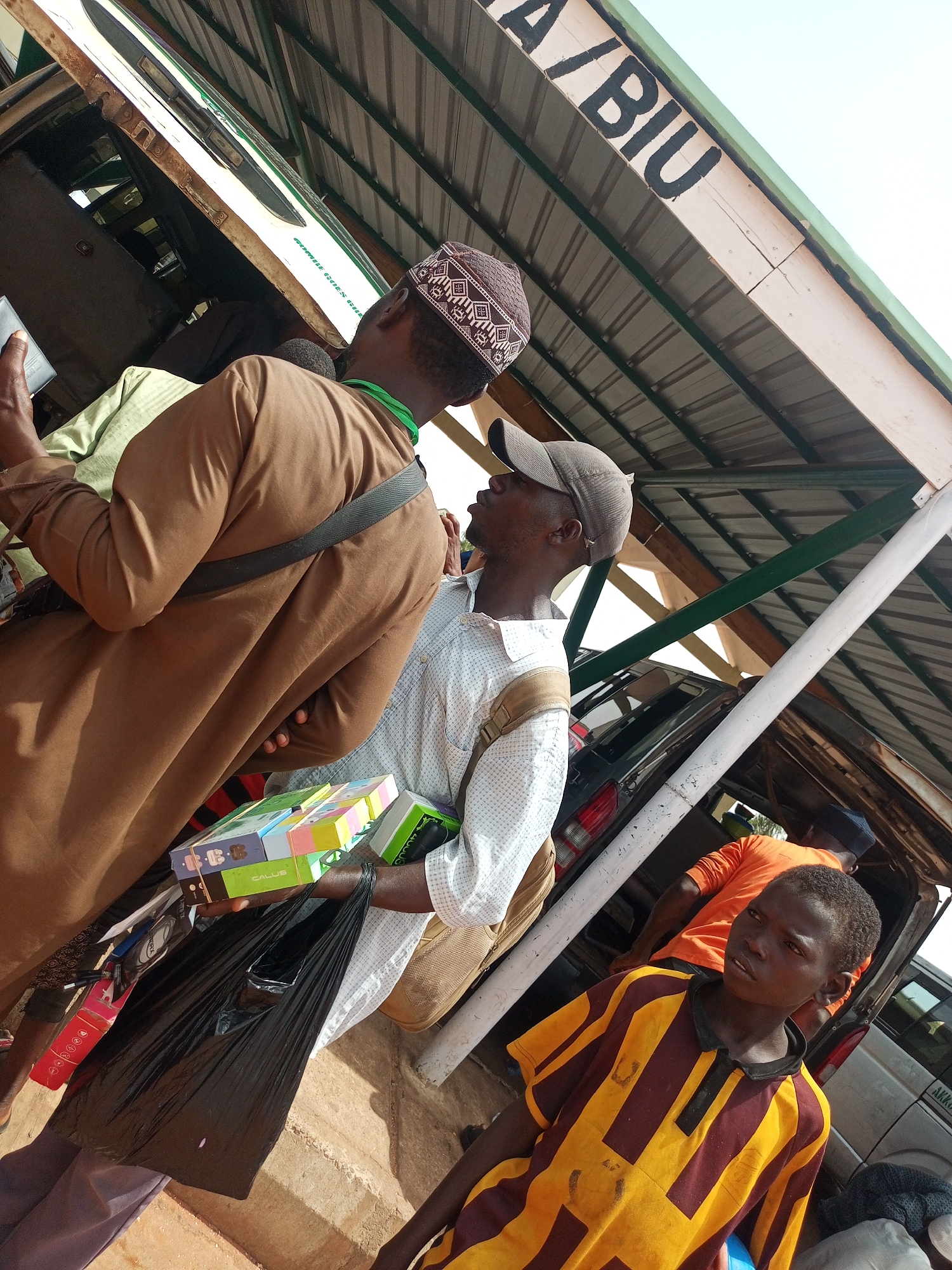 The image shows several people in what appears to be an outdoor market or transportation hub. The location is identified by a sign overhead as "A/BIU" in the city of Unknown, Unknown.
A man in a brown robe and a traditional patterned cap is standing to the left, with a green strap around his neck and holding what looks like a mobile phone. Next to him, another man wearing a gray cap and a white patterned shirt is holding a stack of colorful boxes and a black plastic bag. A young boy in a yellow and brown striped shirt stands in the foreground, looking to the side. In the background, another man is wearing an orange shirt.
Several cars are parked under a metal roof structure.