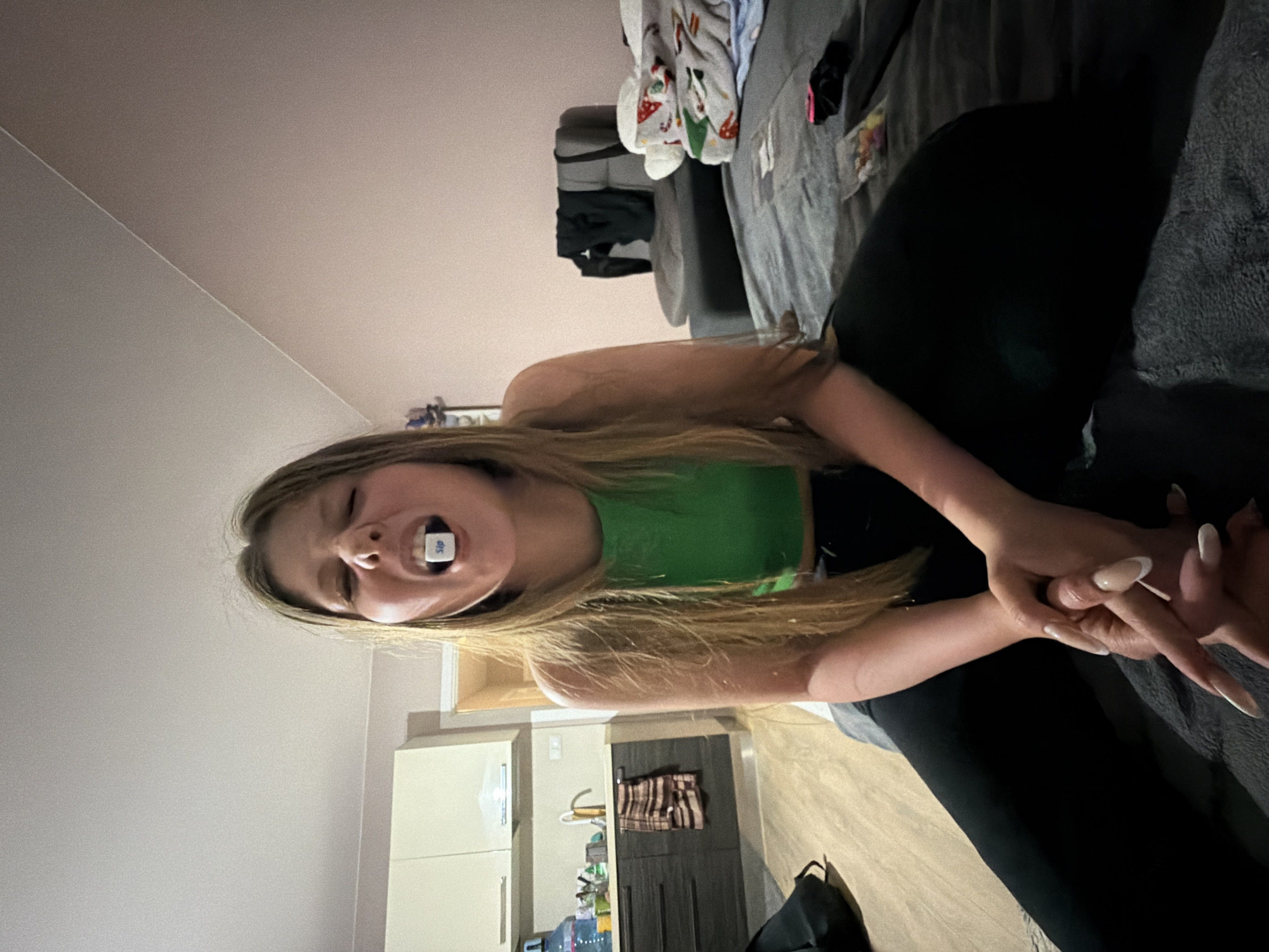 The image depicts a young female in an indoor setting, identified as the city of Burgas, Bulgaria. The individual, possessing long light brown hair, wears a green crop top and dark pants, and is seated on a grey upholstered surface, likely a bed or sofa. Her hands are clasped in front of her. Her mouth is open, holding a white cube with the word "Sip" printed in blue between her front teeth, while her eyes are squinted, indicating a strenuous action or expression.
To the left of the frame, a kitchen area is visible, featuring light beige upper cabinets, dark grey lower cabinets, a dark countertop with a silver-faucet sink, and a large blue water dispenser bottle. A striped towel hangs from a cabinet handle. The floor in this area is a light wood-look material, with a black bag positioned on it.
To the right and behind the female, a grey armchair is present, draped with dark clothing. The background wall is a plain light pink color. On the grey upholstered surface, next to the female, a patterned blanket with festive designs is visible, along with a clear plastic bag containing small, colorful objects and other miscellaneous items. The scene is illuminated by interior lighting.