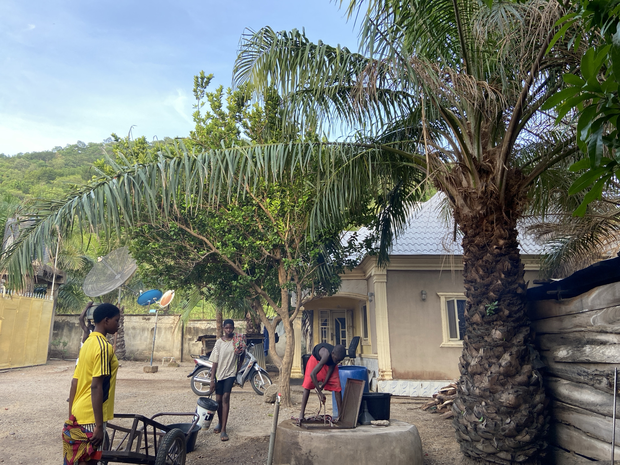 The scene is a yard in Mararaba, Nigeria, displaying daily life activities. In the foreground, a person wearing a yellow shirt and patterned skirt is standing beside a two-wheeled cart. Near them, another individual with a striped shirt carries a bucket. A third person, in a red shirt and shorts, appears to be working on equipment atop a cylindrical well structure. A motorcycle is parked nearby.
The background features a single-story residential building with light-colored walls and a gray tiled roof. Various trees and a large palm tree provide shade. A satellite dish is visible on a pole. The ground is dirt and gravel. In the distance, a hill or mountain covered in vegetation can be seen.