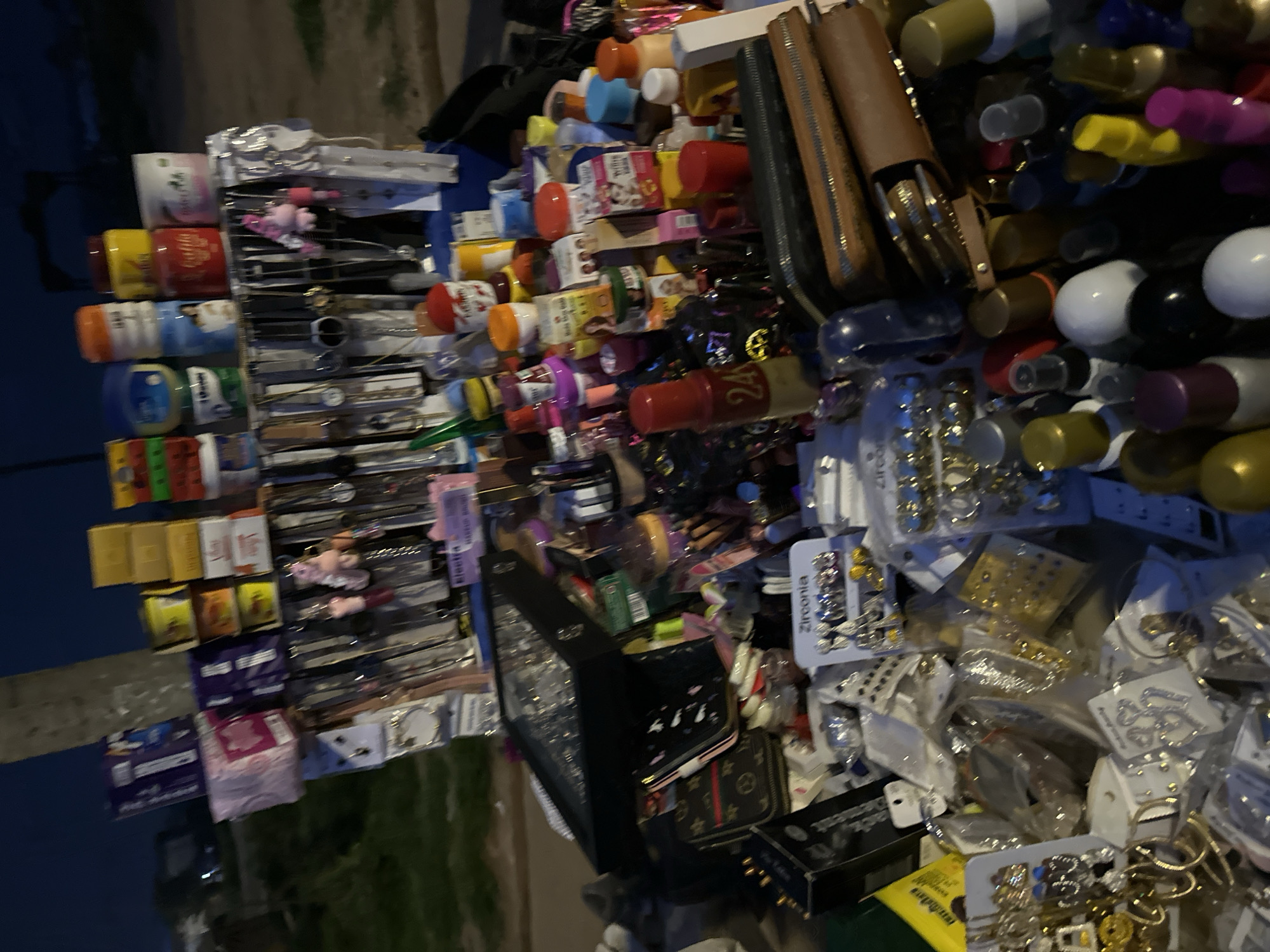 The image depicts a street vendor stall in Bukuru, Nigeria, at dusk. The stall is laden with various merchandise, including an assortment of watches, necklaces, earrings, and other fashion accessories displayed on racks and scattered across the surface. Several jars of cosmetic products, such as petroleum jelly and lotions, are also visible. A black jewelry box with a clear lid is open, revealing more accessories. The vendor appears to be absent from the frame. The background shows a dimly lit street with a pole and some foliage, suggesting an outdoor market setting. The ground is covered with dirt and debris.
