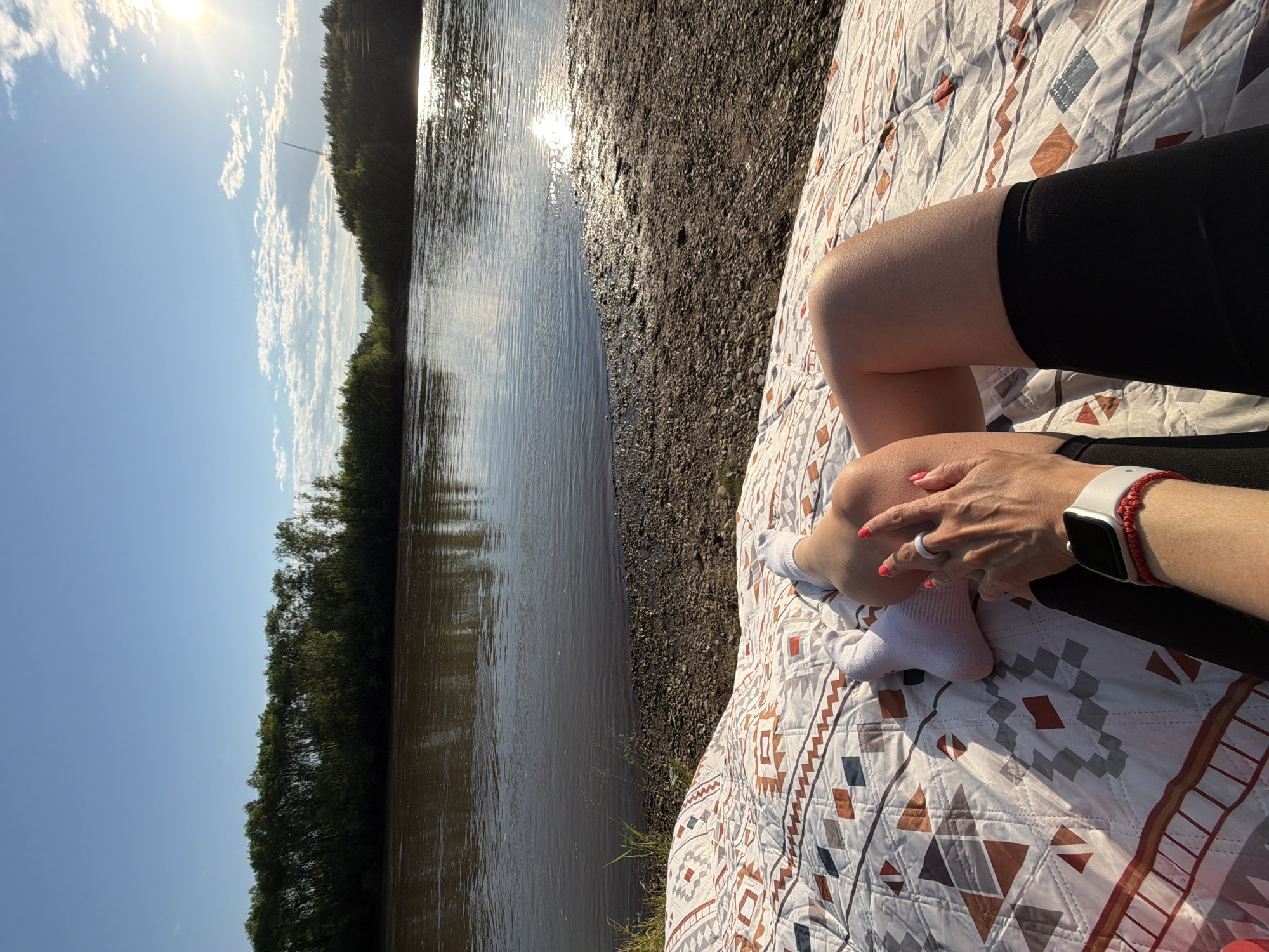 The image depicts a first-person perspective of someone relaxing on a blanket near a river. The blanket is laid out on a pebbly shore, and the river is wide with calm water reflecting the sky and trees. A dense line of trees borders the far bank of the river. The sky is a clear blue with scattered white clouds, and the sun is setting or rising, casting a bright glare on the water's surface. The person's legs are visible, wearing white socks and black athletic shorts. One hand is resting near their legs, showing a white smartwatch and a red bracelet. The nails are painted bright pink. The setting appears to be natural and tranquil, possibly a park or recreational area. The location is stated to be Birobidzhan, Russia.