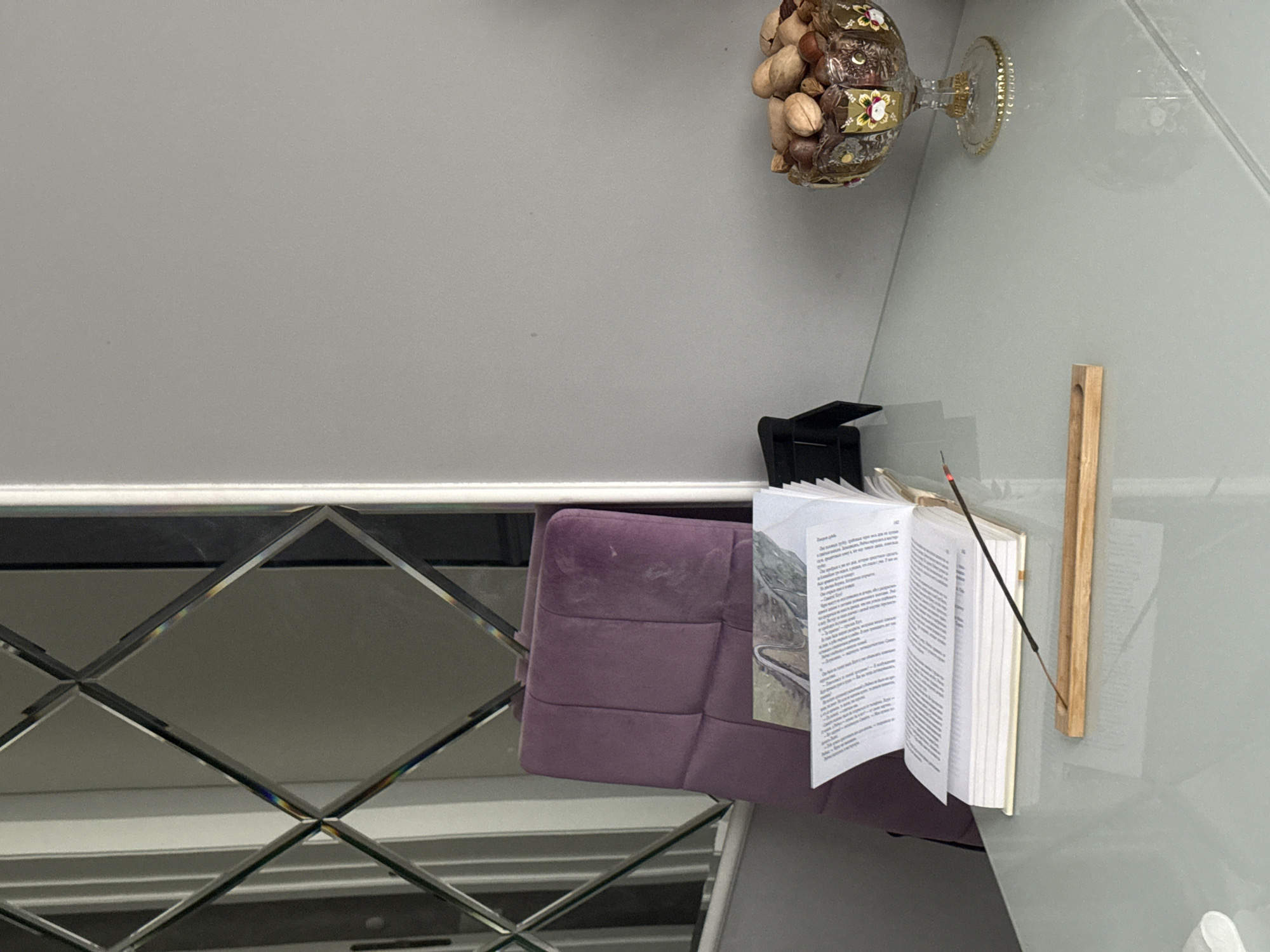 A low-angle shot captures an interior scene with a reflective, diamond-patterned surface in the foreground. Above this, a purple, cushioned object rests, seemingly attached to a wall or panel. An open book stands atop this cushioned object, its pages filled with text and a landscape image visible on one side. An incense stick is placed within the book's pages. To the right of the book, a slender wooden object is affixed to the wall. In the upper right corner, a clear glass container with nuts rests on a shelf. In the upper-left, a gray wall provides a backdrop. The location is Unknown, Unknown.