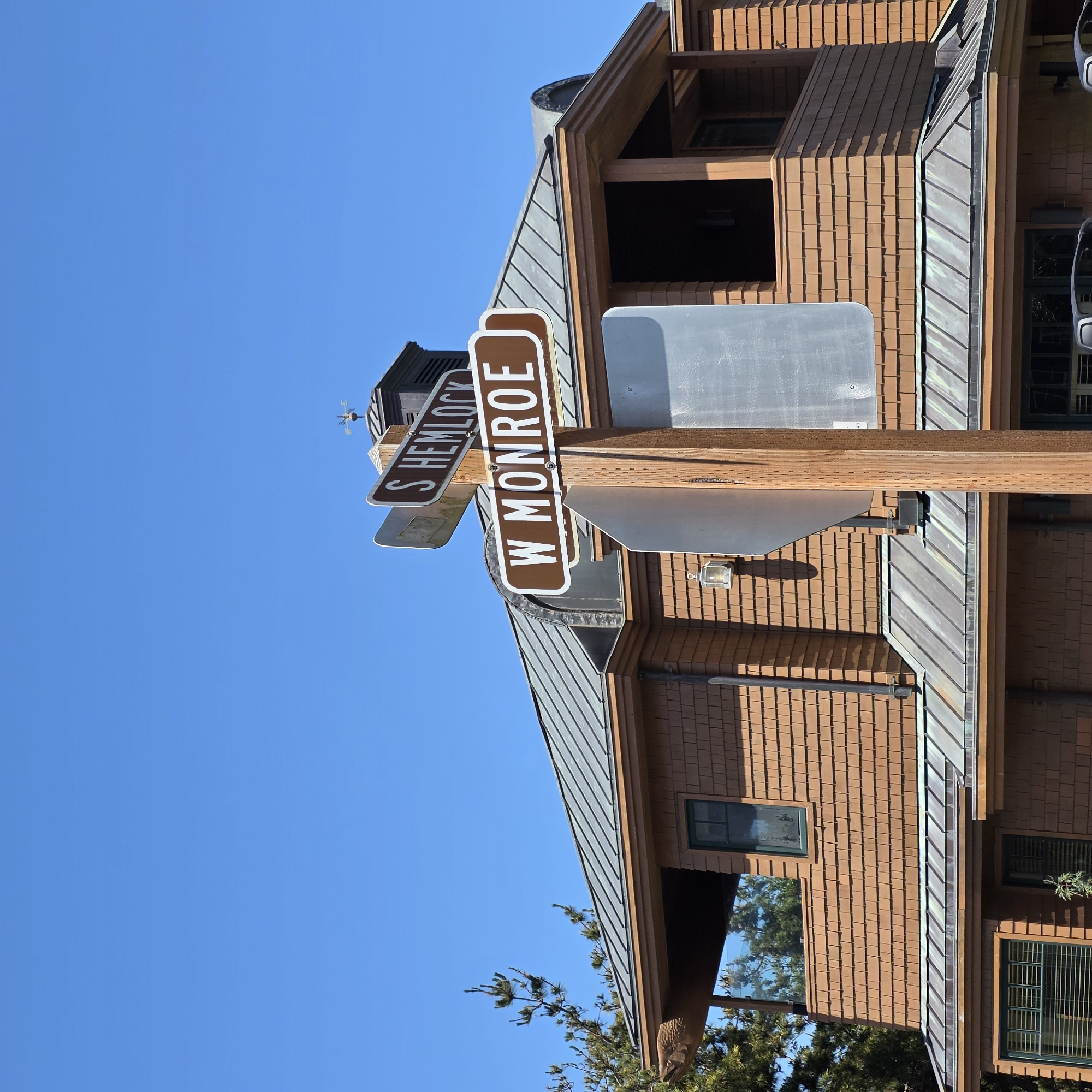 In Cannon Beach, Oregon, United States of America, a two-story wooden house with a shingled exterior is shown on a clear, blue sky day. A prominent wooden pole stands in front of the house, supporting two brown street signs with white lettering. The upper sign reads "S HEMLOCK" and the lower sign reads "W MONROE." To the left of the pole, a portion of the house's roof is visible, which appears to be made of dark gray metal. A small, decorative wooden fish carving protrudes from the eaves of the roof on the left side of the image. A weather vane is visible on top of a dark cupola on the roof in the background. The house has several windows with dark green frames. Trees with green foliage are present on the left side of the image.