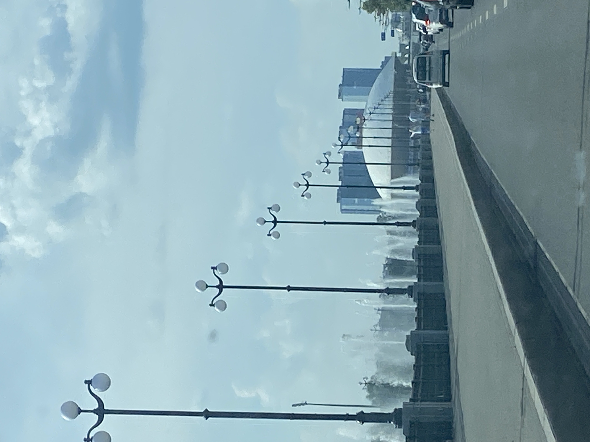 The image depicts a cityscape under a partly cloudy sky. In the foreground, a paved road leads towards a row of cars in traffic. The road is bordered by a gray curb and sidewalk. Along the sidewalk, a series of ornate streetlights with three globes each are evenly spaced. Behind the sidewalk, a dark stone wall with decorative elements runs parallel to the road, and behind this wall, a large fountain with multiple jets of water is visible. In the background, several modern buildings with glass facades rise into the sky, some appearing as residential complexes and others as office buildings. A distinctive white dome-shaped structure is situated among these buildings, with more streetlights extending towards it. The scene suggests a public space, possibly a waterfront promenade or a grand avenue in a city. Based on the visual cues of the architecture and the presence of the fountain and streetlights, this scene is likely located on Kremlevskaya Embankment in Kazan, Russia. There are a few individuals walking on the sidewalk in the middle ground, and traffic is moving along the road in the foreground.