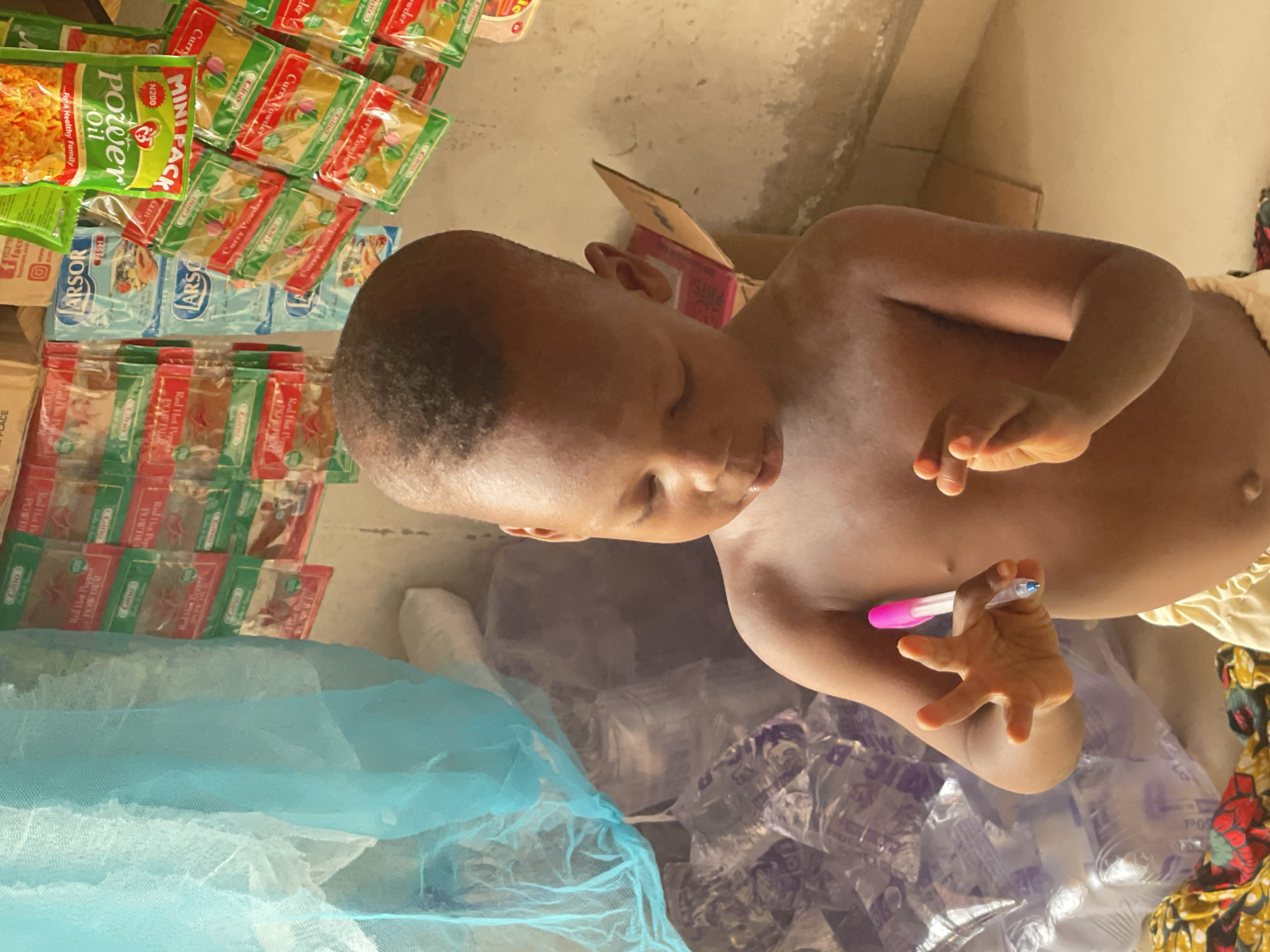 The image, rotated 90 degrees clockwise, shows a young child holding a pink-capped pen. The child is shirtless and wears light-colored bottoms, looking down with a focused expression. In the background, shelves or stacks are filled with various packaged food products. Notable items include "Gino" brand "Curry Powder" and "Red Hot Pepper Powder," as well as "Power Oil" (100% Refined Palm Olein) with the slogan "...For A Healthy Family." One "MINI PACK" of "Power Oil" is priced at "N200," indicating Nigerian currency. Other packages are labeled "ARSOR" and "HST." A light blue, net-like fabric rests in the lower left foreground, and clear plastic bags are visible underneath. The setting appears to be an indoor space, likely a small shop or storage area, consistent with a commercial or domestic environment in Lokoja, Nigeria. The bright, even lighting suggests it is daytime.