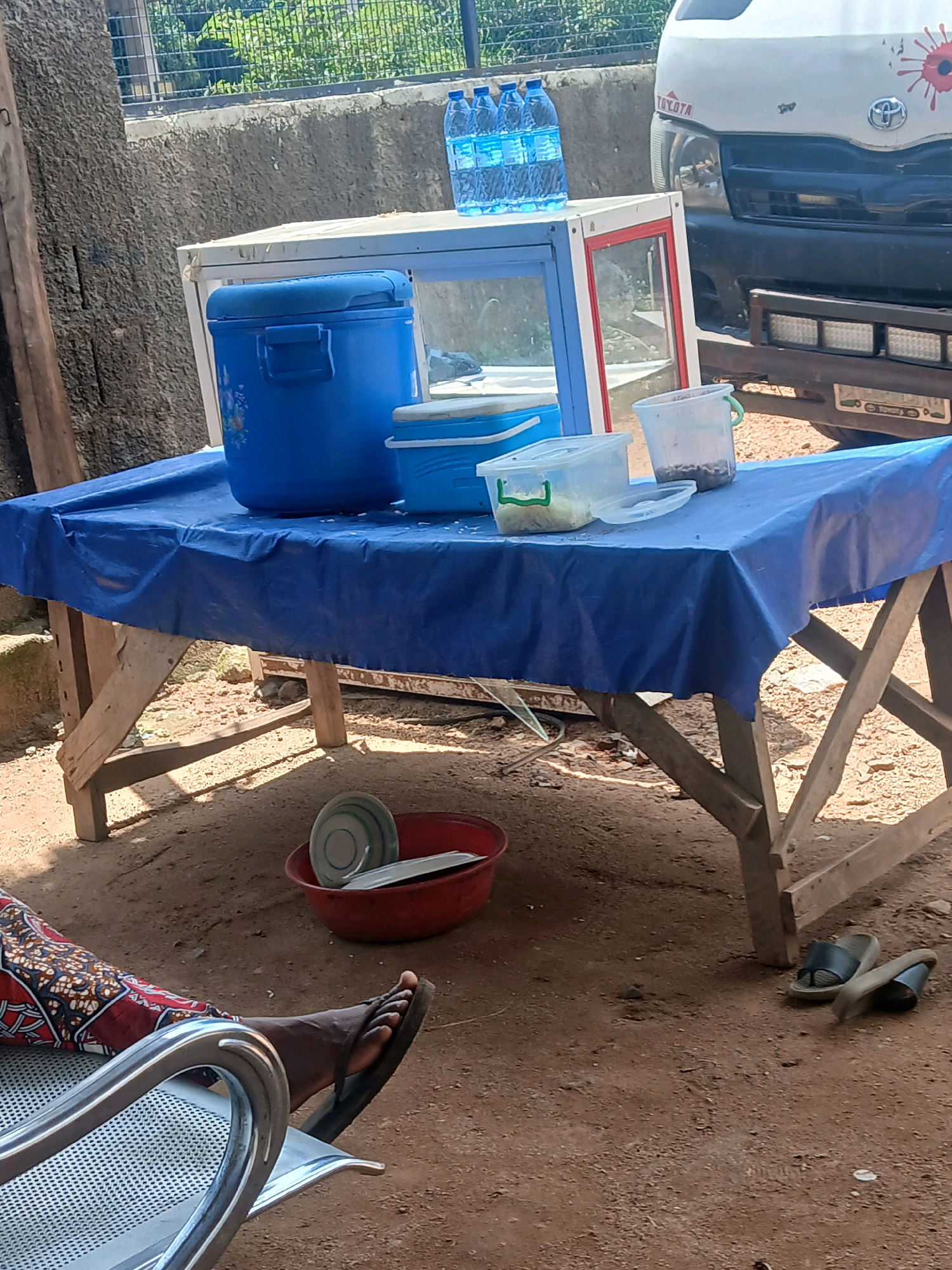 In a dirt-floored outdoor setting, likely a roadside stall, a wooden table draped with a blue plastic tarp supports various items. Prominently featured is a large blue cooler with a handle. Adjacent to it are two smaller blue containers and two clear plastic containers, one holding what appears to be food items and the other holding a dark substance. On the table, behind these containers, are several bottles of water. To the right of the table, the front of a white Toyota van is visible. On the ground near the table, a red plastic basin contains stacked plates and cutlery. A person is seated in a metallic chair to the left, with their legs and feet visible, wearing a patterned cloth and flip-flops. Another pair of flip-flops rests on the ground to the right of the table. The background consists of a textured wall and foliage behind a fence.