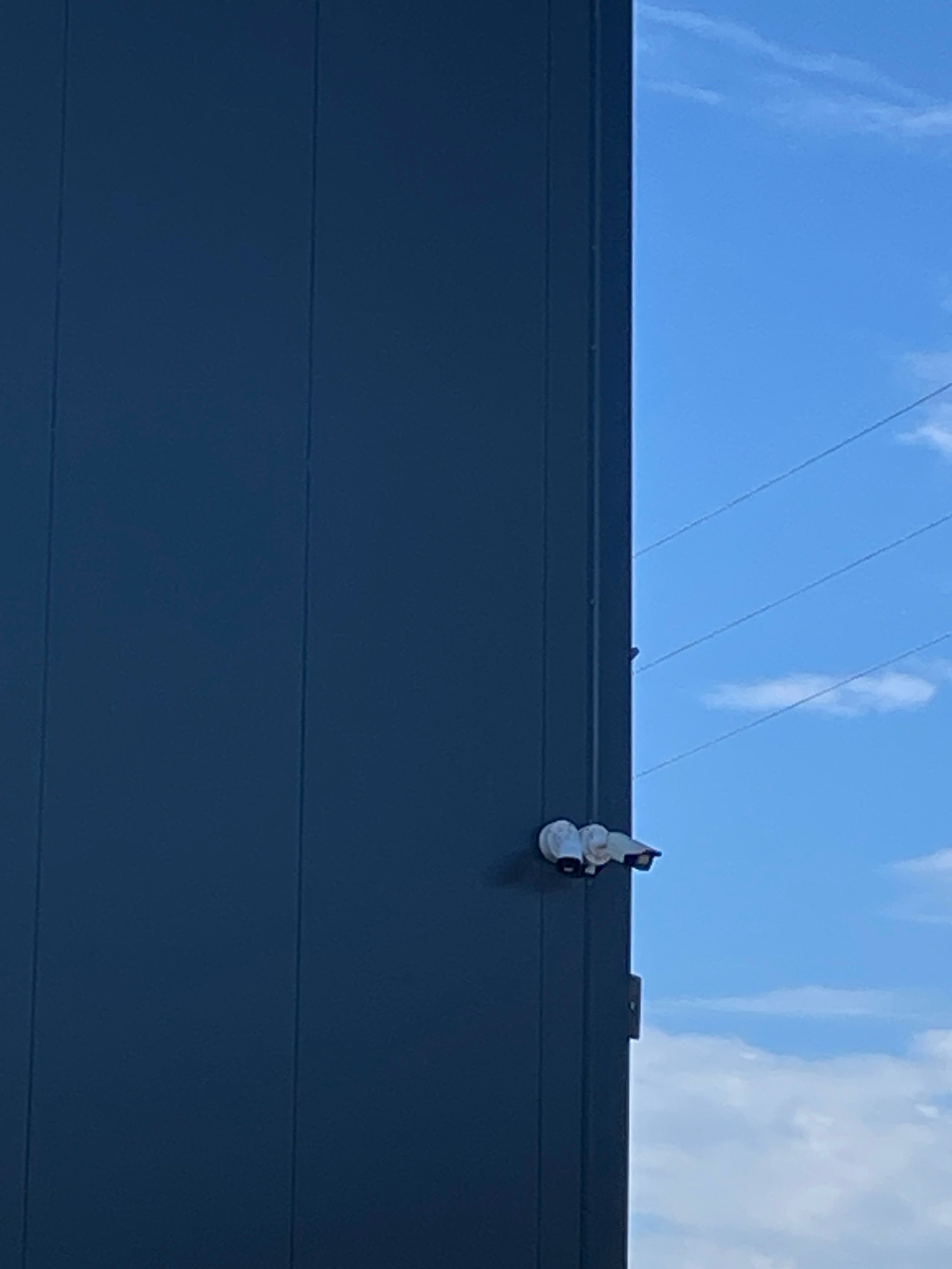 An outdoor, medium-angle, eye-level shot shows the side of a dark blue building with a bright blue sky and some white clouds in the background. The building takes up the left two-thirds of the image and has vertical seams running from top to bottom. Two white security cameras are mounted on the right side of the building. The cameras are positioned close to each other, with one camera slightly above and to the left of the other. Power lines can be seen stretched across the sky in the upper right portion of the image.