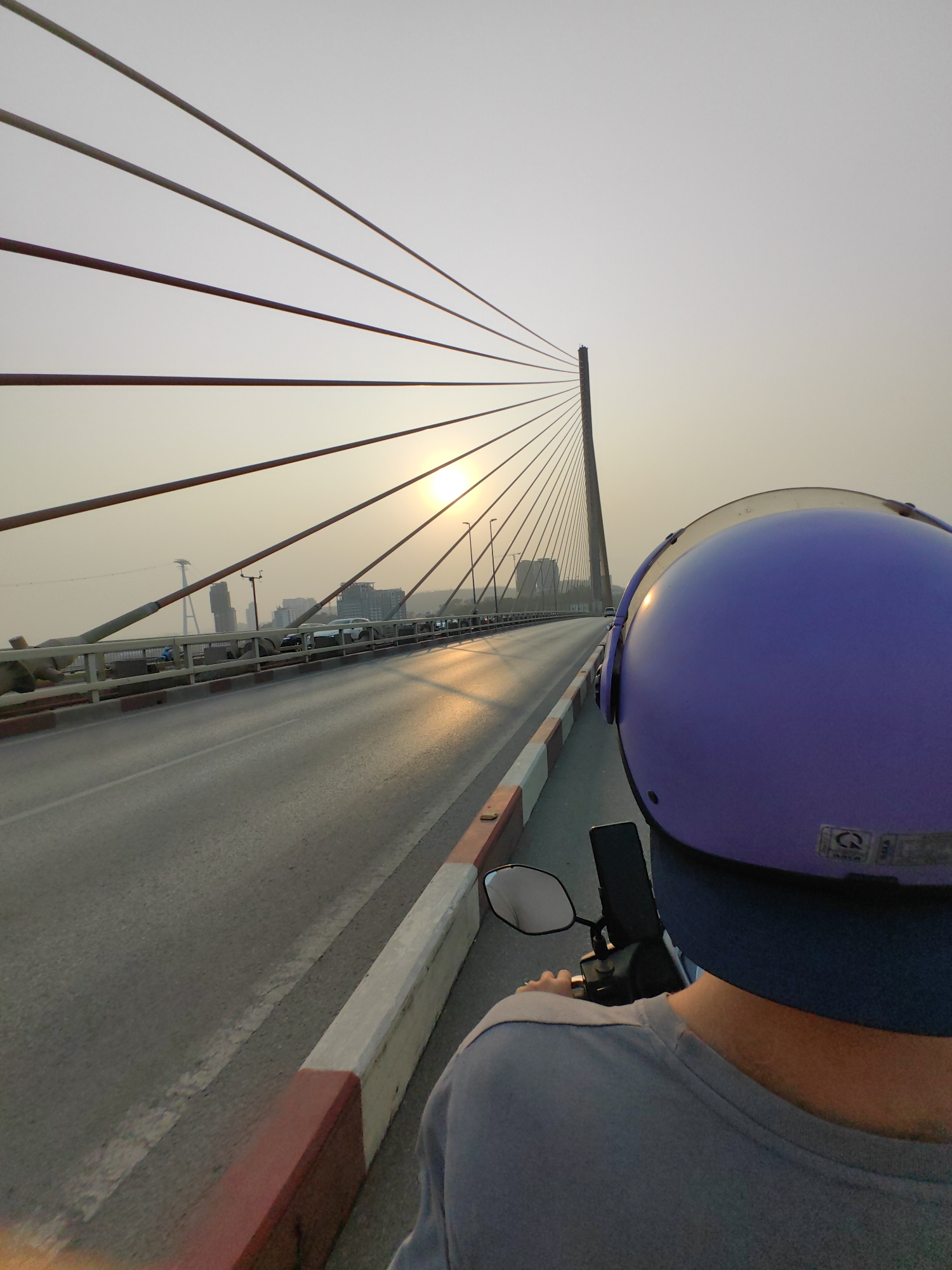 View from the back of a person riding a motorcycle on Bai Chay Bridge, Hong Gai Ward, Vietnam, at sunset. The person is wearing a purple helmet. The bridge's cable stays are visible. The sun is positioned near the bridge's tower, causing a glare. Other vehicles and buildings are visible in the distance.