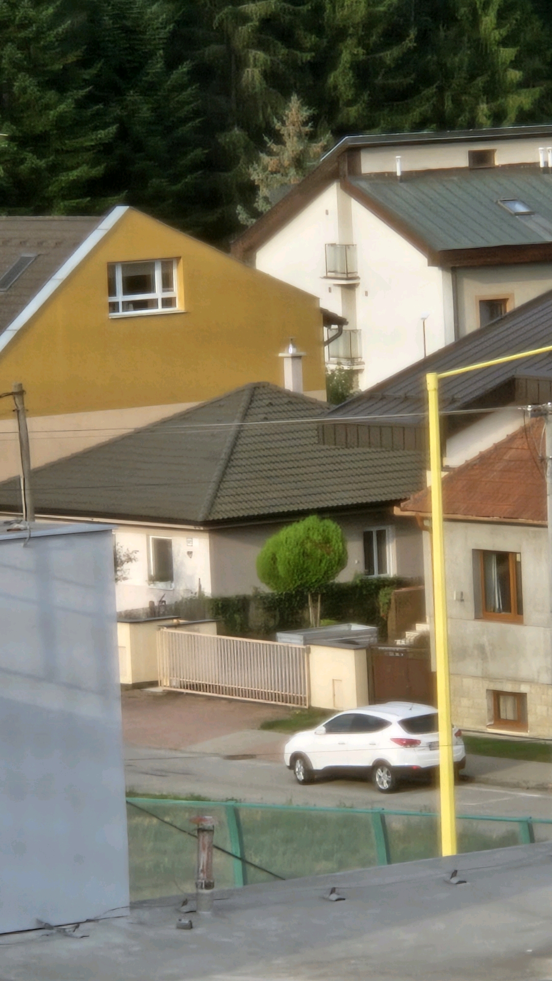 The image depicts a residential area with multiple houses and a dense forest in the background. The architecture suggests a European setting, likely a suburban or semi-rural part of Prešov, Slovakia, given the context provided. The foreground shows a grey flat rooftop with some exposed piping and what appears to be a vent. A green-paneled fence is visible. In the mid-ground, a white SUV is parked on a paved driveway next to a building with a dark tiled roof and a bright orange-tiled roof on an adjacent structure. A tall yellow pole stands near the buildings. A beige fence with vertical slats separates the driveway from a small yard with a neatly trimmed green shrub. Further back, a yellow-painted house with a single white-framed window is visible, nestled against a larger white building with a dark green roof. The background is dominated by a lush green forest, characteristic of the natural landscapes surrounding Prešov. No people are visible in the image.