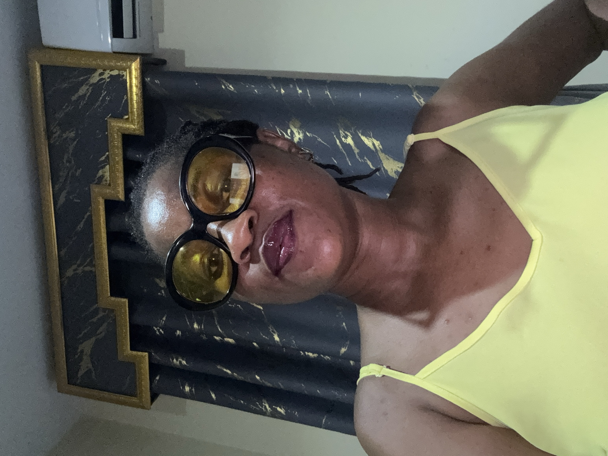 In the city of Ajara, Nigeria, a medium shot captures a Black woman wearing large, round, yellow-tinted sunglasses. She is adorned in a bright yellow spaghetti-strap top. Her lips are painted with a dark, glossy lipstick. Her dark hair is styled in braids, with some strands visible around her face. The background features dark blue curtains with a golden, abstract pattern, and a decorative gold and black valance above them. To the right of the frame, a portion of a white air conditioning unit is visible against a light-colored wall.