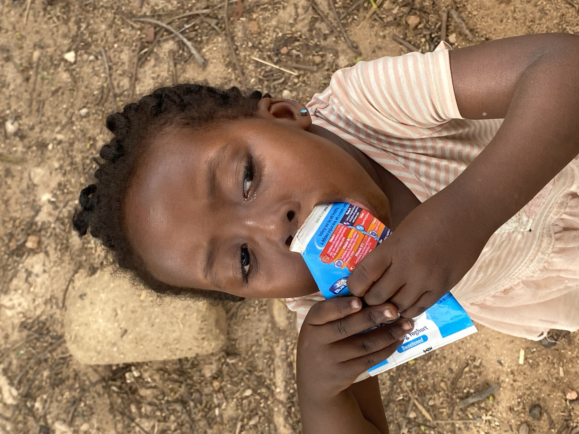 The image depicts a young Black child drinking from a "Blue P" brand yogurt drink. The child, wearing a light pink striped top and a matching dress, is positioned on what appears to be dry, cracked earth with small stones and twigs scattered around. The yogurt drink packaging is white and blue with nutritional information printed on it. The background suggests an outdoor setting, possibly in a rural or impoverished area. The photo was taken in Kuru Dungus, Nigeria.