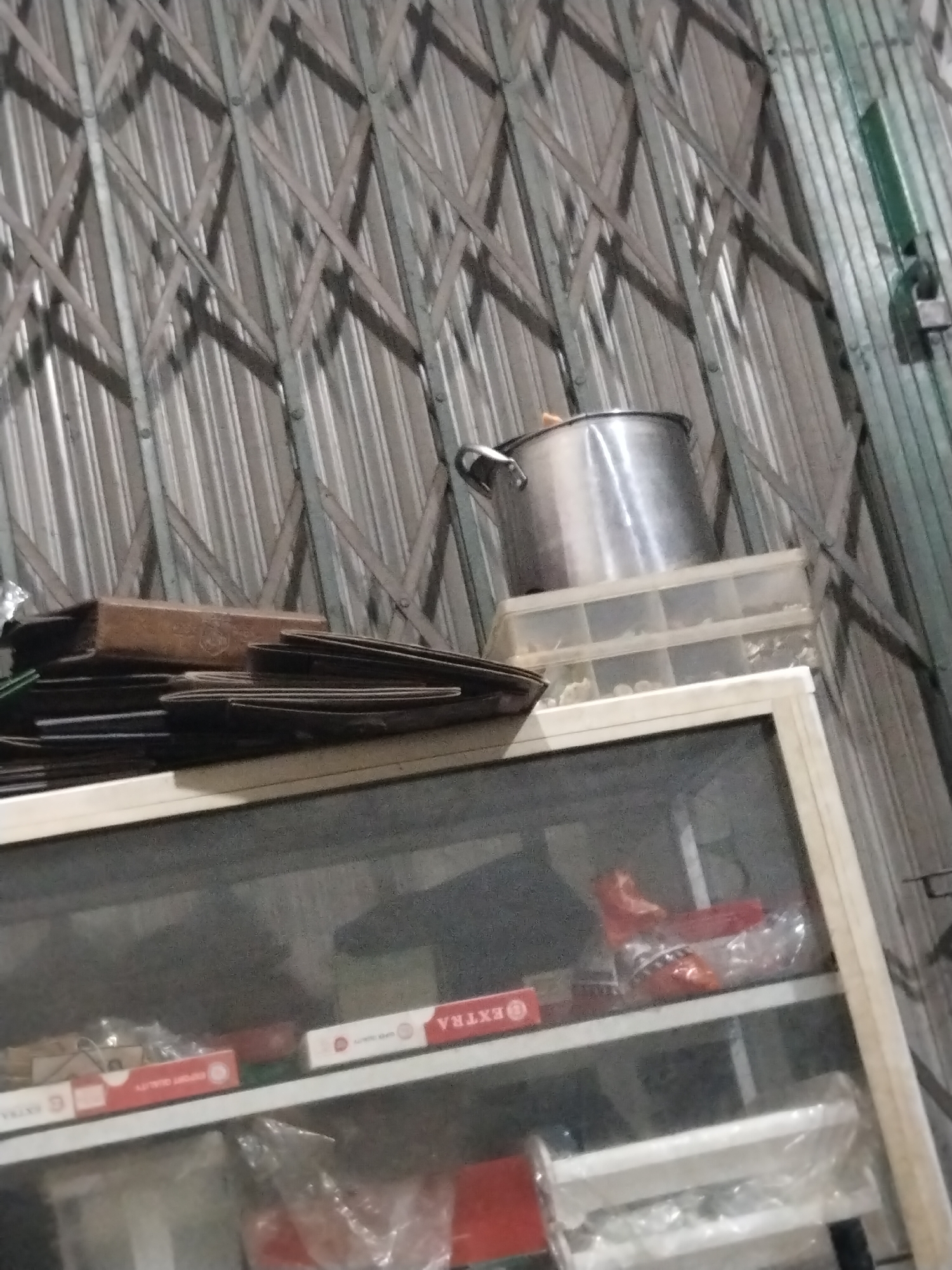 The image shows a cluttered shelf against a corrugated metal wall with a retractable gate. On the top shelf, there is a large stainless steel pot on top of a clear plastic organizer box. To the left of the pot, there is a stack of what appears to be cardboard boxes or flat objects. Below this, the shelf holds several items including red and white boxes with text, one of which reads "EXTRA" and "SUPER QUALITY". There are also plastic-wrapped items on this shelf. The bottom shelf is mostly obscured but contains more plastic-wrapped items. A green metal structure with a lock is visible on the right side of the image, attached to the wall. The lighting is dim, casting shadows across the scene. The overall impression is of a storage area or a workshop.