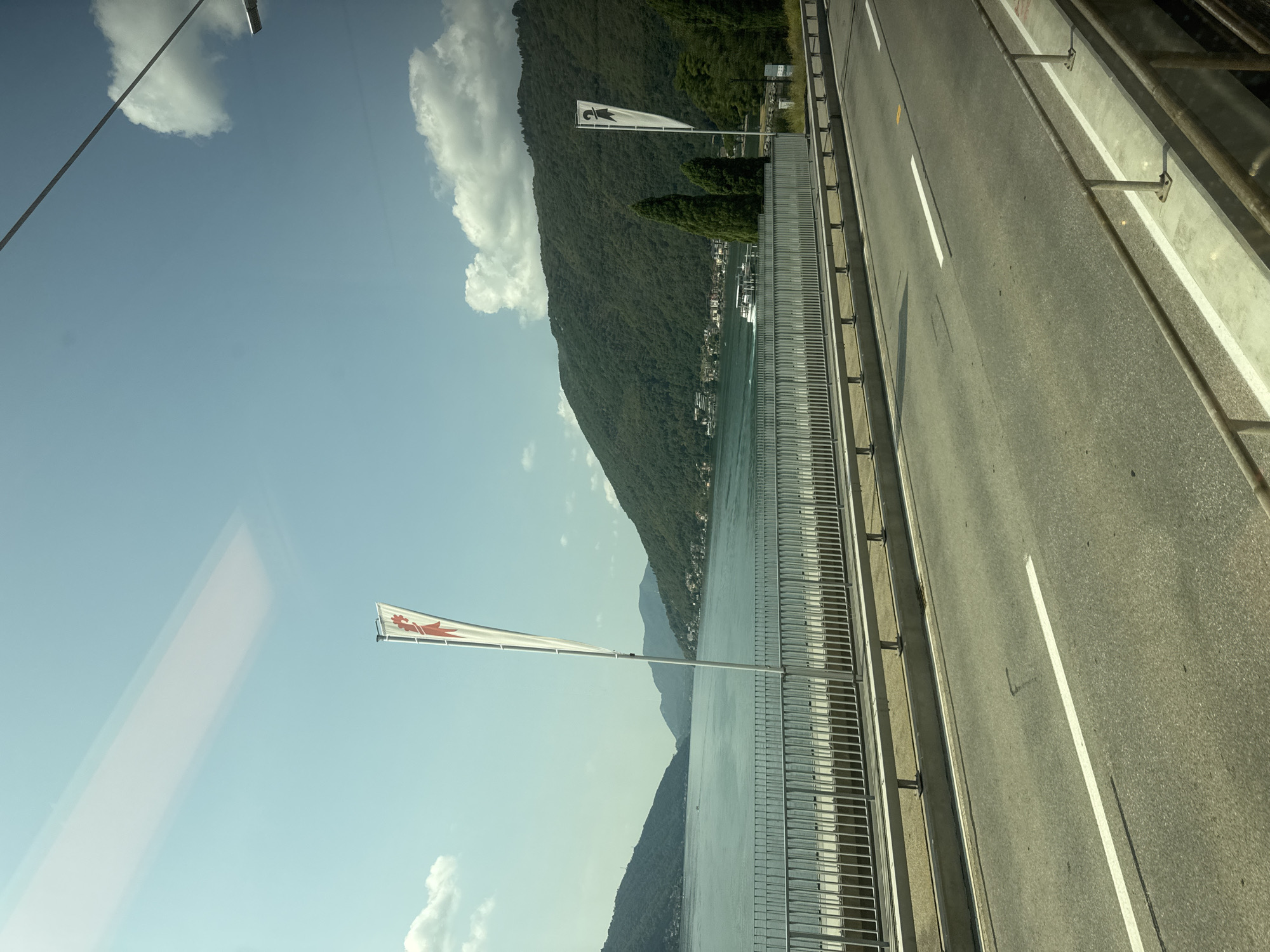 The image shows a view of Lake Lugano from a road or bridge in Bissone, Switzerland. On the left, there is a white flag with a red graphic, possibly a bird or rooster. In the background, green, tree-covered mountains rise from the lake. A boat is visible on the water in the distance. The road in the foreground has white lane markings. A metal railing runs along the edge of the road, separating it from the lake. The sky is blue with scattered white clouds.