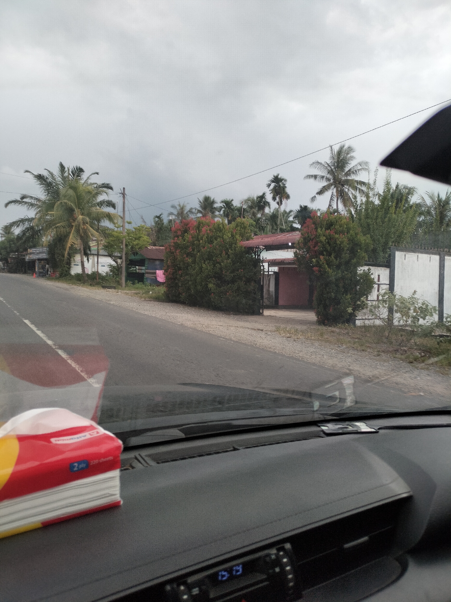 A car's dashboard is in the foreground, with a packet of tissues resting on it. Beyond the car's windshield, a road is visible, lined with a white dashed line on the left side. On the right side of the road, there is a fence with white panels and dark borders. Behind the fence, houses and lush green vegetation, including palm trees and flowering bushes, are present. The sky is overcast with gray clouds. The scene appears to be on a road in Bukit Selamat, Indonesia.