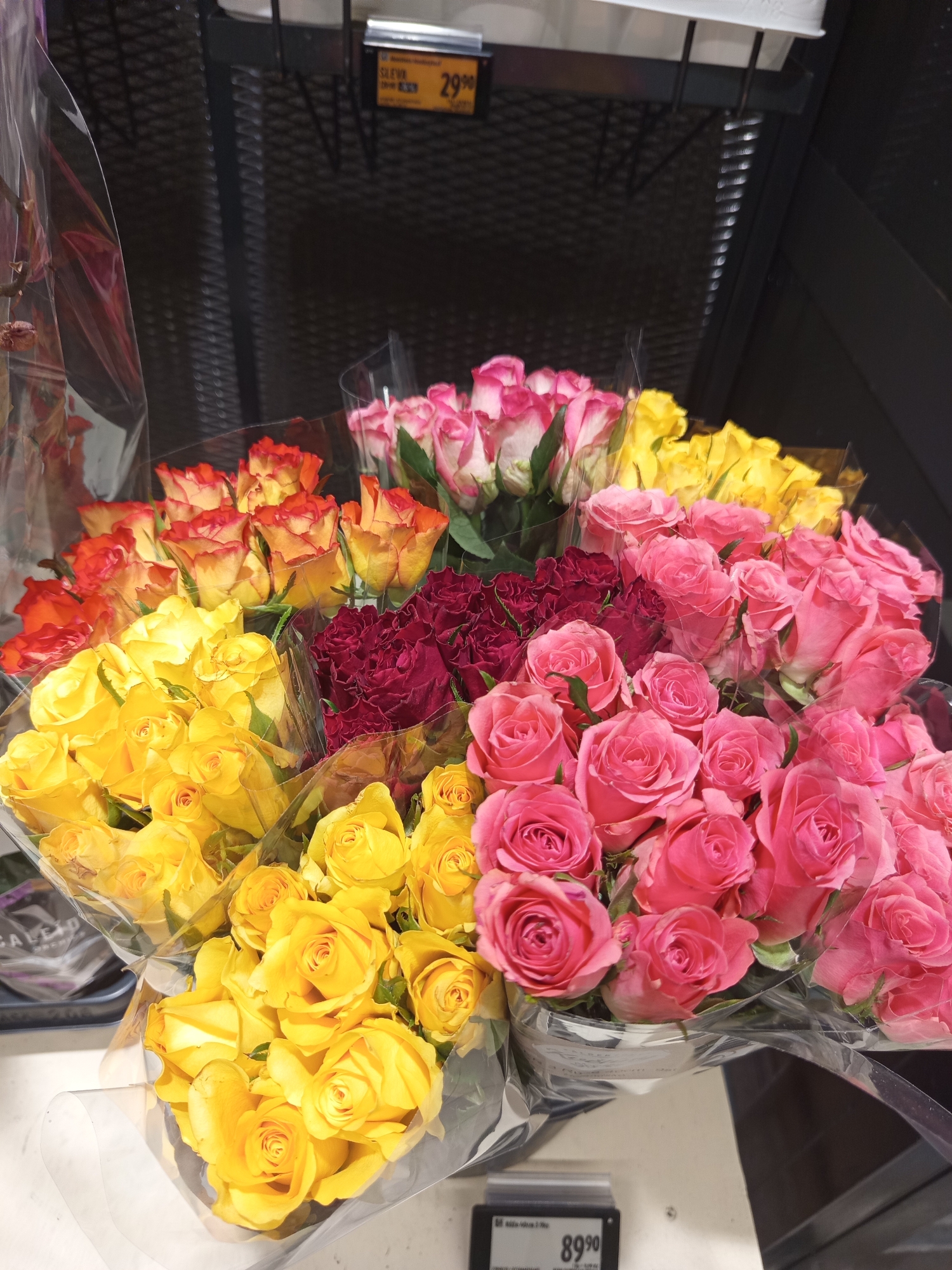 The image depicts a display of bouquets of roses in a store. The roses are arranged in several bunches, with varying colors including yellow, pink, red, and bi-colored orange and yellow. The bouquets are wrapped in clear plastic. In the background, a price tag displaying "29.90" and another with "89.90" are visible. The overall scene suggests a retail environment, likely a flower shop or a supermarket floral section. There are no people visible in the image. The lighting is bright, indicating an indoor setting. It is not possible to determine the specific location within Brno, Czechia from the visual information provided.