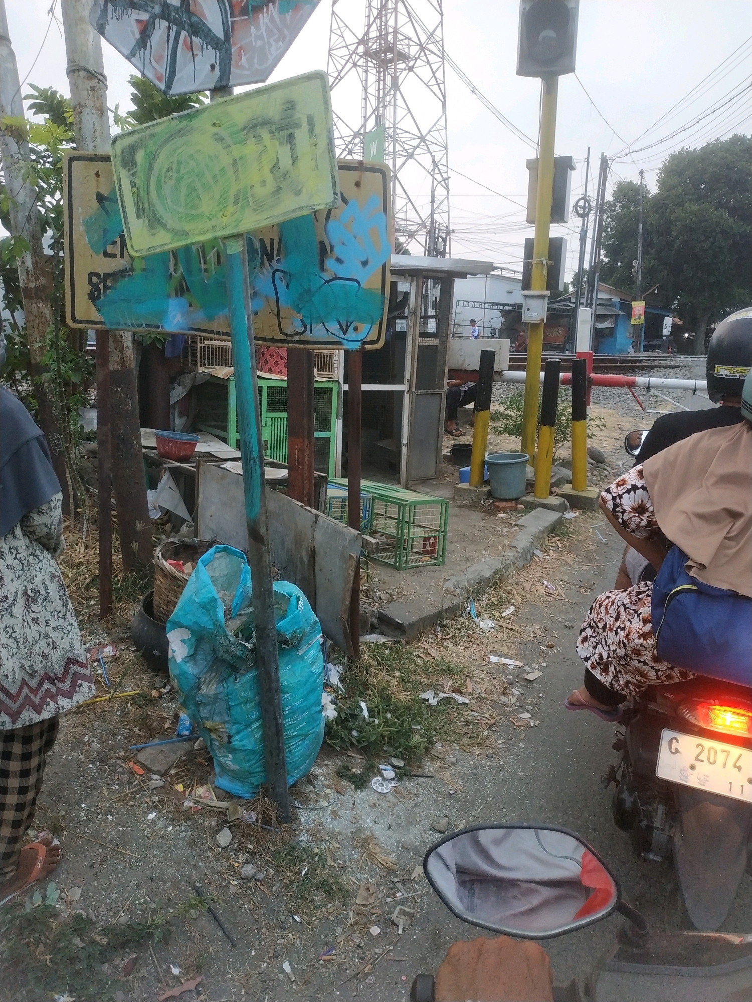 In Tegal, Indonesia, a photograph captures a roadside scene near a railway crossing. A person, partially visible on the left, wears a patterned garment and an orange sandal, standing near a metal pole with a yellow sign covered in graffiti. Behind this, a turquoise plastic bag lies on the ground amidst debris, near a woven basket. The background features a railway line with a crossing barrier, a signal post, and a red and white striped structure. A motorcycle with a license plate reading "G 2074 11" is positioned to the right, with two individuals seated on it. One person, wearing a black helmet and a light brown head covering, is on the back. The environment appears somewhat dishevelled, with scattered trash and vegetation.