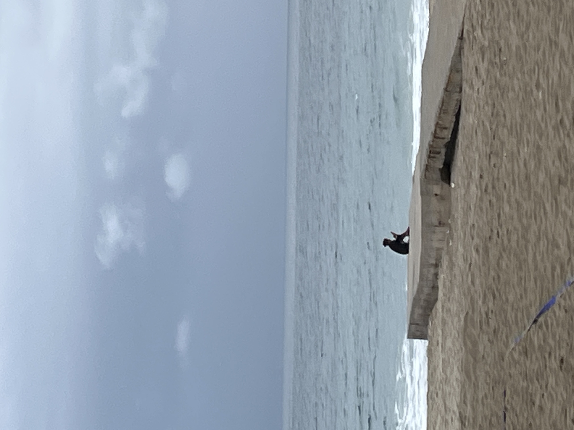 The image shows a beach scene with a concrete structure separating the sand from the water. A dark figure is standing on the concrete, next to the water. The sky is overcast with clouds. It is possible that this is Alanya, Turkey, due to the type of beach and structure but I cannot confirm this.