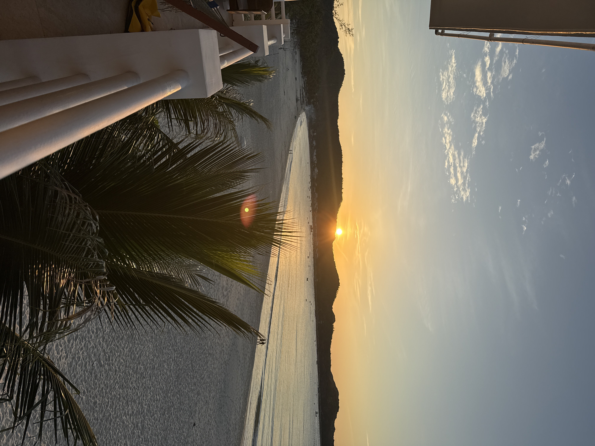 This is a photograph of a beach at sunset, taken from an elevated perspective, likely a balcony. The sun, a bright orb, is setting behind a line of hills, casting a warm, golden glow across the sky and the calm water. The beach itself is sandy, with a gentle curve. In the distance, along the shoreline, there are small boats and structures that suggest a settlement. Two figures are walking along the water's edge, silhouetted against the bright sea. In the foreground, the fronds of a palm tree obscure part of the view, and to the left, a white railing and the corner of a building are visible. The sky is a gradient of pale blue at the top, transitioning to a soft orange and yellow near the horizon, with scattered wispy clouds. The overall scene depicts a tranquil coastal landscape at dusk.