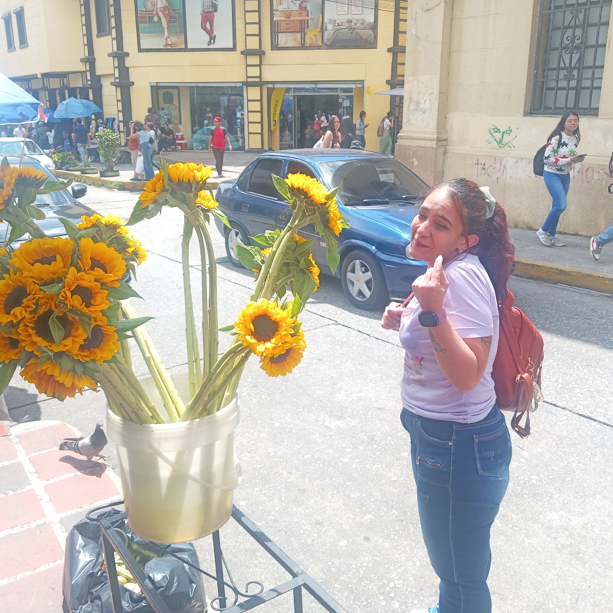 The image depicts a street scene with a woman standing next to a bucket of sunflowers. The woman is wearing a light purple t-shirt, blue jeans, a red backpack, and a smartwatch. She is smiling and pointing with her right index finger.
Next to her is a bucket filled with sunflowers, placed on a metal stand. There is a black garbage bag hanging below the bucket. A pigeon stands on the sidewalk beside the sunflowers.
Behind the woman and the sunflowers, a blue car is parked on the street. In the background, there is a yellow building with multiple floors, windows, and what appear to be advertisements. People are walking in the background, both on the sidewalk and entering/exiting the building. Graffiti can be seen on the wall of a building to the right.