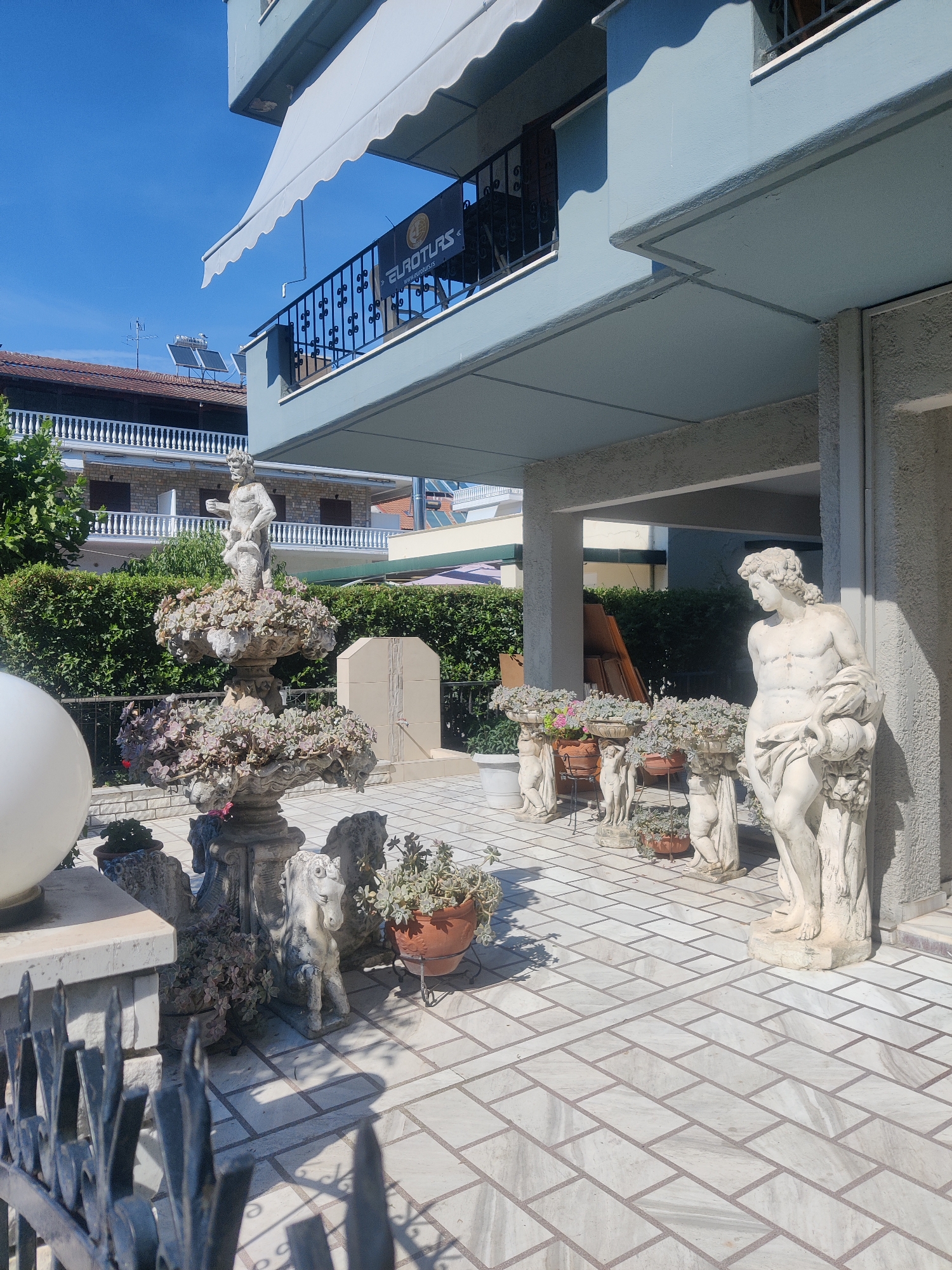 The image shows an outdoor patio area decorated with classical-style sculptures and potted plants. A multi-tiered fountain with a statue on top is visible in the center-left of the frame, surrounded by succulent plants. To the right, a statue of a male figure in a draped garment stands near a pillar. Several smaller statues of cherubs and horses are also present, along with various potted plants. The patio floor is tiled with light-colored rectangular stones. In the background, a blue building with a balcony and an awning is partially visible, along with neighboring structures and greenery. The scene appears to be a residential or commercial property in a sunny outdoor environment. The specific location of Katerini Municipality, Greece is not evident from the visual content of the image itself.