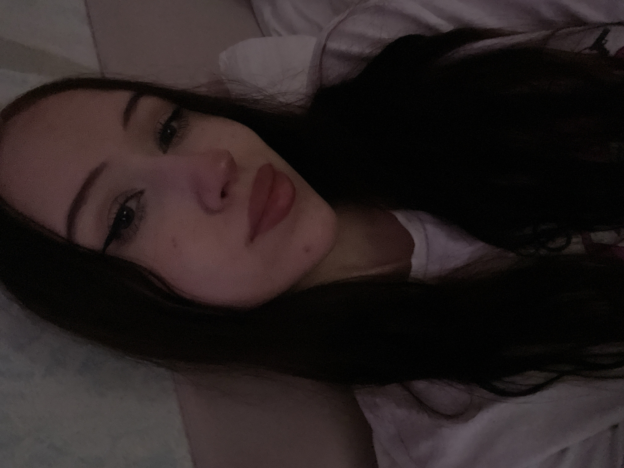 The media file is a close-up, vertically oriented selfie of a young person with long, dark hair. The subject is lying down, likely in a bed, as indicated by the soft, textured surface beneath them which appears to be a pillow or blanket. The lighting is dim and appears to be artificial, suggesting it may be nighttime or indoors with low light.
The subject's face is angled towards the camera, with their eyes open and looking directly at the viewer. They have dark eyelashes and eyebrows, and their lips are slightly parted, with a subtle sheen suggesting lip gloss or balm. A hint of eyeliner is visible on their upper eyelids. The overall mood conveyed is intimate and possibly relaxed, given the reclining pose.
There are no discernible objects other than the subject and their immediate surroundings, which appear to be a bedding environment. No text is visible in the image. The provided location context of Votkinsk, Russia, does not offer any specific visual cues within the image itself.