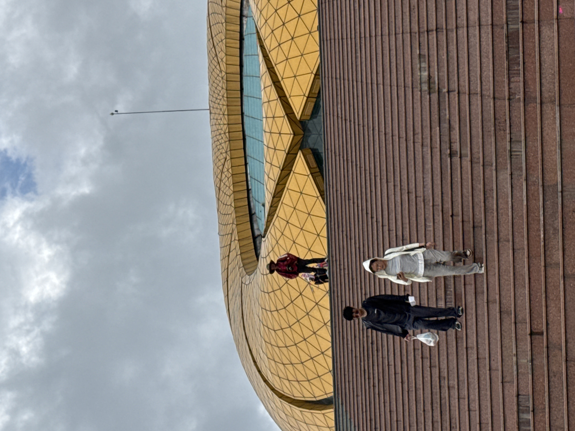 The image depicts a modern architectural scene in Da Lat, Vietnam, under a heavily overcast sky. A striking golden building, characterized by a complex, multi-faceted facade of geometric, likely triangular, panels, dominates the left side of the frame. The building features distinct curved sections and reflective blue-tinted glass windows. A tall, thin pole, possibly a flag mast or antenna, extends upwards from the building against the backdrop of the grey clouds.
In front of the golden structure, a wide expanse of dark reddish-brown steps or a tiered plaza stretches across the foreground and middle ground. These steps have a distinct ridged texture and show some minor discolorations or wear.
Three individuals are visible on these steps, appearing to walk casually across the scene. In the immediate foreground, a person wearing a black jacket, dark trousers, and possibly a dark hat or dark hair, carries a white plastic bag in their left hand. Behind them and slightly to the left, another person is dressed in a white hooded jacket, light grey pants, and light-colored shoes. Further back, closer to the base of the golden building, a third person is seen wearing a reddish or burgundy patterned long-sleeved shirt and a hat.
The cloudy sky indicates a daytime setting, though direct sunlight is absent. No specific emotions are discernible from the individuals, and there is no visible text in the image.