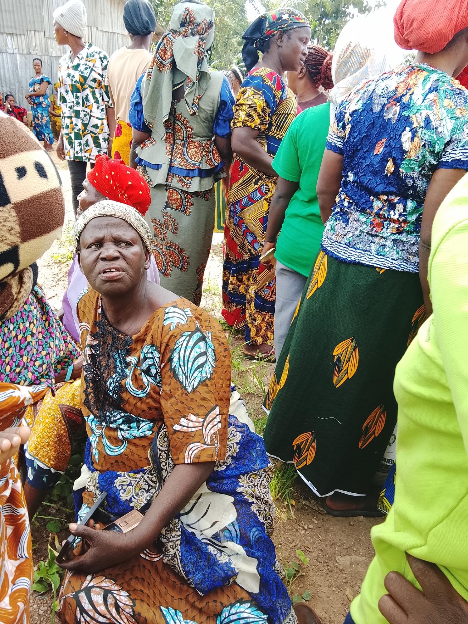The image shows a group of people, primarily women, gathered outdoors in what appears to be a rural or semi-rural setting in Akwanga, Nigeria. The lighting suggests it is daytime, likely morning or afternoon, with diffused light indicating possible cloud cover. The ground is a mix of dirt and sparse vegetation, with a building structure visible in the background, possibly a home or community building with a corrugated metal roof.
The subjects are dressed in colorful, patterned clothing, common in West African attire. Many are wearing headscarves or headwraps. One woman in the foreground, seated and facing the camera, is wearing a brown and blue patterned dress and a light-colored headscarf. She has a concerned or serious expression. Another woman in the middle ground, also seated, is wearing a red headscarf and a patterned dress. Several other individuals are visible, mostly from the shoulders up or in profile, also in brightly colored clothing.
The women are gathered together, but the specific activity is unclear. It could be a social gathering, a community meeting, or people waiting for an event. One woman in the foreground is holding a dark-colored flip phone in her hand. There is no visible text in the image. The overall mood seems to be one of community and everyday life, with the vibrant clothing providing a striking contrast to the simple, earthy surroundings.