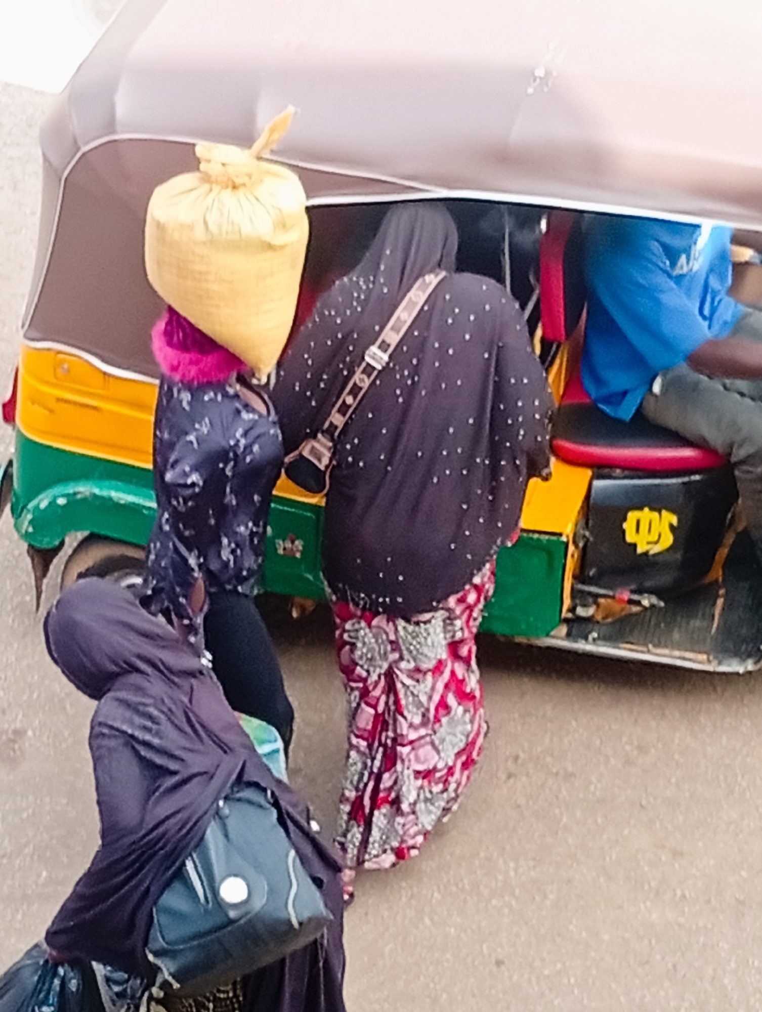 In Bukuru, Nigeria, an auto-rickshaw is loaded with passengers and goods. A woman in a purple headscarf and dark clothing, carrying a large dark bag on her shoulder, stands to the left of the frame. To her right, another woman, seen from behind, wears a dark, speckled abaya and a bright red and white patterned skirt, and has a yellow bag tied to her head. A third person, partially visible, is seated inside the auto-rickshaw, wearing a blue shirt. The auto-rickshaw itself is brightly colored with yellow, green, and black sections. The scene appears to be taking place on a paved road.
