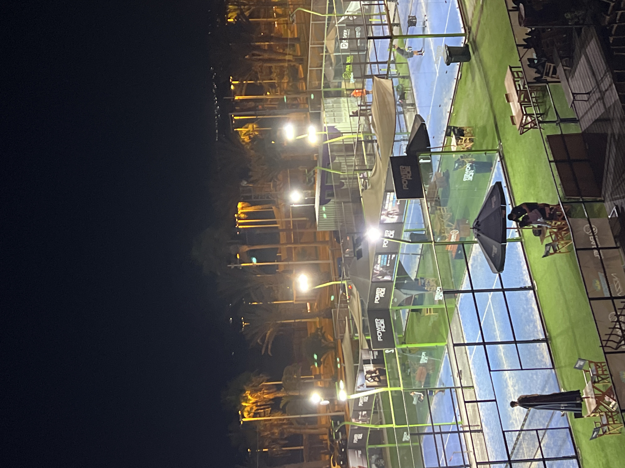 The image shows a nighttime view of multiple padel tennis courts. The courts are illuminated, revealing blue playing surfaces and green surrounding areas. Glass walls and metal frames enclose each court. People can be seen playing and spectating. Several branded advertising signs surround the courts, with "POWERADE" prominently displayed. Palm trees and streetlights line the perimeter. The background is dark, suggesting a late hour. There is no visual evidence to specifically identify the location as Arica, Chile.