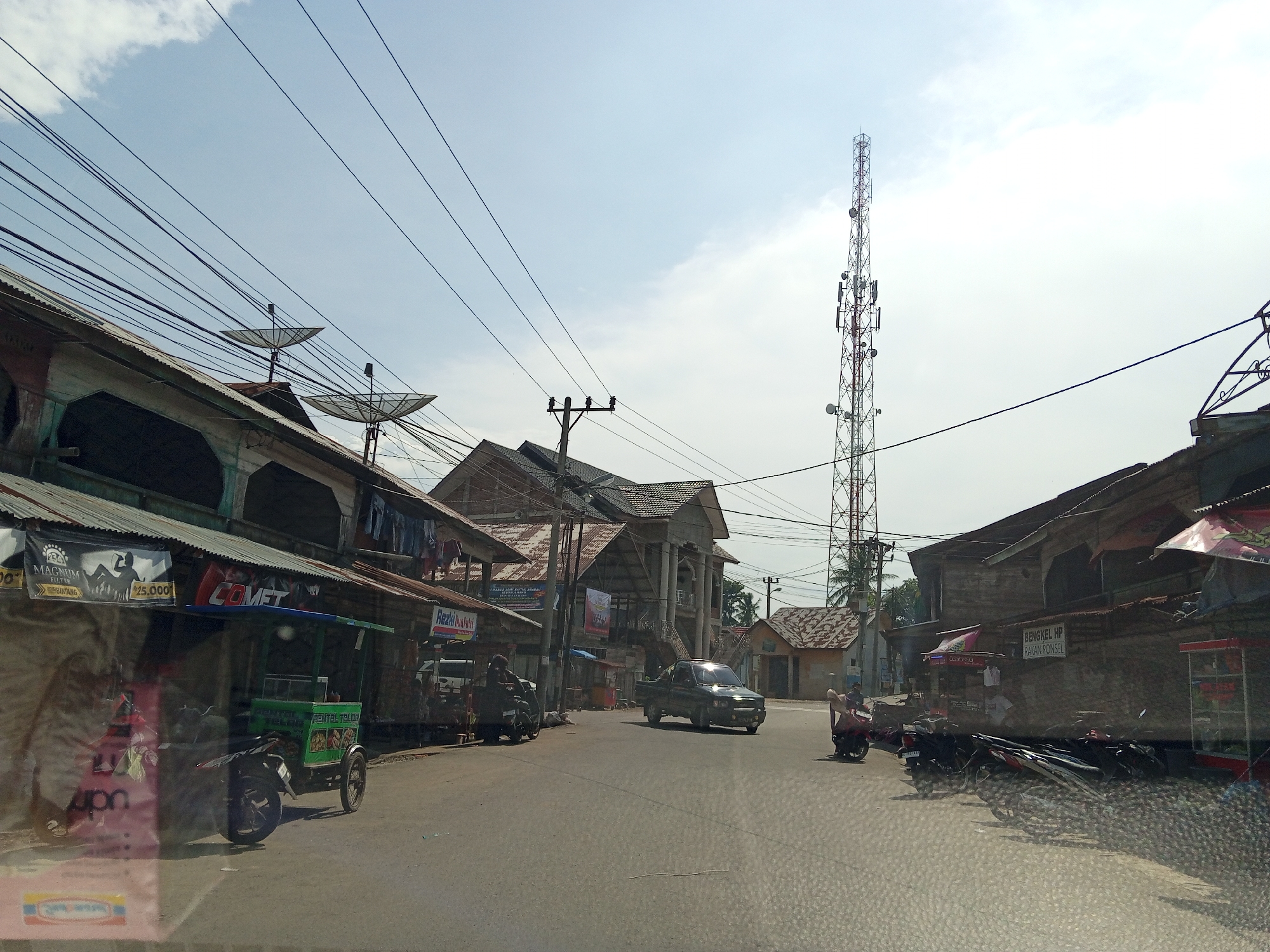 This image depicts a street scene in Meuriya, Indonesia, under a clear to partly cloudy sky. The perspective is from a vehicle, with minor reflections visible in the lower foreground.
The paved road occupies the lower and central portion of the frame, showing faint tire marks and surface texture. A dark-colored pickup truck drives centrally, facing the viewer, with its headlights glinting in the sunlight. To the right of the pickup, a person on a red scooter with a passenger is visible, positioned near the curb. Further right, several motorcycles are parked along the side of the road next to a building.
On the left side of the street, a line of weathered, single and multi-story buildings with corrugated metal roofs runs parallel to the road. Multiple utility poles with a dense network of overhead wires crisscross the sky. Several satellite dishes are mounted on the rooftops of these buildings. Commercial banners and signs are affixed to the building facades, including "MAGNUM FILTER," "COMET," and a green food stall attached to a motorcycle, with "PENTOL TELOR" visible on its side. Another motorcycle is parked further down this side of the street.
In the mid-ground, a larger, two-story building with a more ornate facade, featuring columns and external staircases, stands behind the initial row of structures on the left.
On the right side of the street, similar weathered buildings with corrugated roofs are present. A tall, red and white cellular/telecommunication tower, equipped with multiple antennas and dishes, dominates the right-hand sky, extending above the rooftops. Palm trees and other foliage are visible in the background, behind the buildings and the tower.