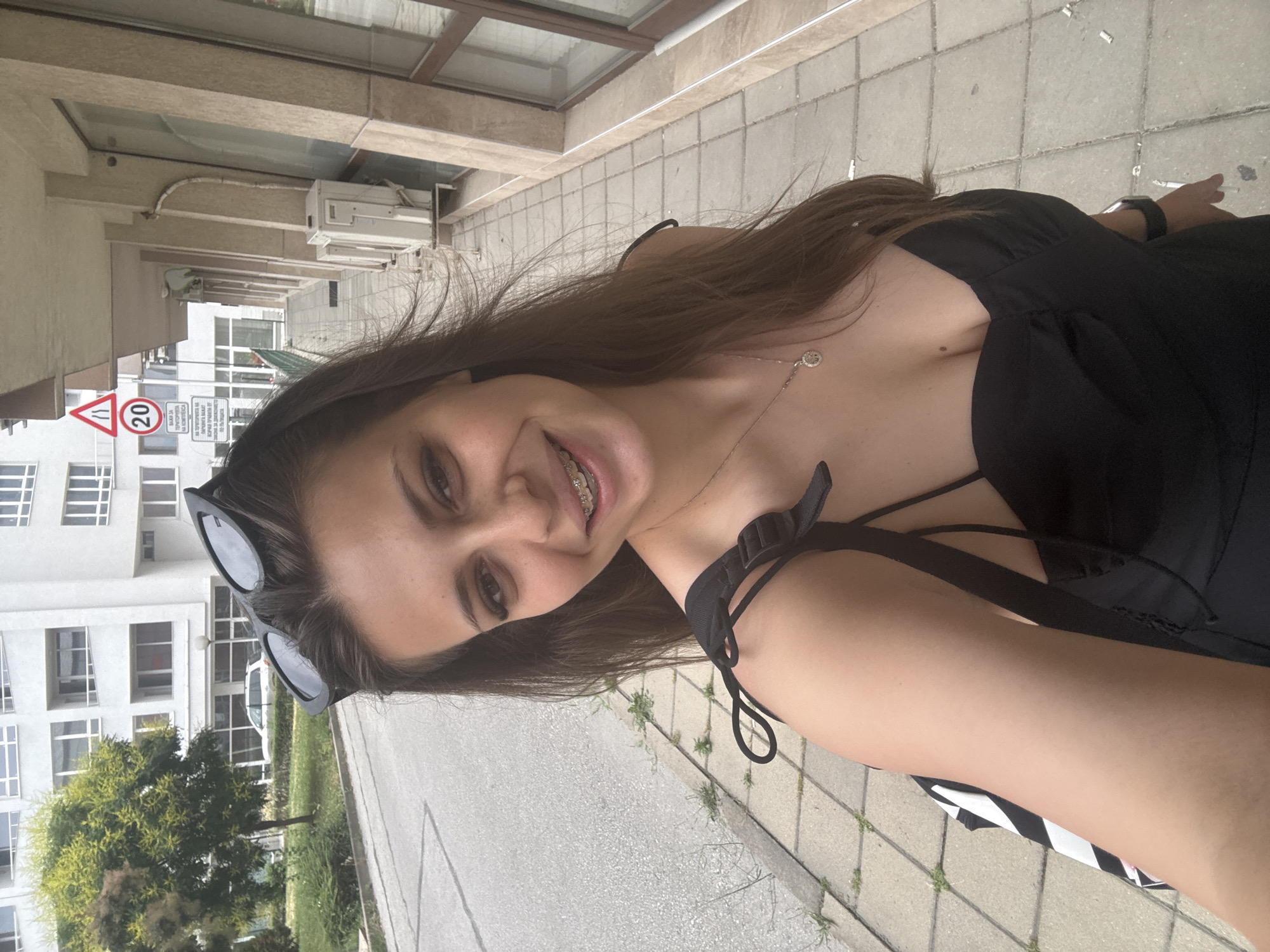 A young woman with long brown hair, braces, and sunglasses perched on her head is taking a selfie outdoors. She is wearing a black dress with thin straps and a pendant necklace. Behind her, a white building with balconies is visible, along with a paved sidewalk and a street. Traffic signs, including one indicating a 20 km/h speed limit, are posted near the building. The scene appears to be in a residential or commercial area of Plovdiv, Bulgaria, suggested by the architecture and the presence of signs in Bulgarian.