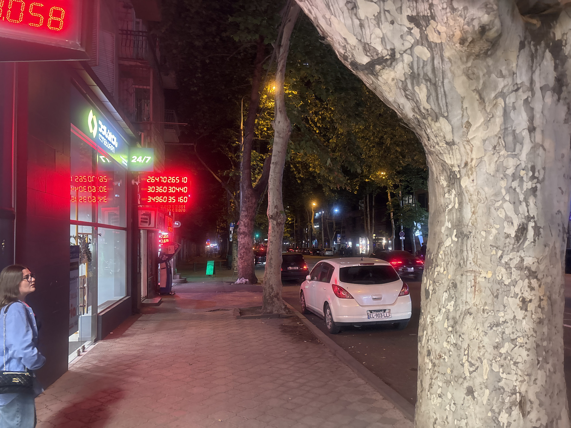 The image depicts a street scene at night in Tbilisi, Georgia, with a prominent digital exchange rate display showing currency conversions. The display exhibits rates for USD to GEL, EUR to GEL, and GBP to GEL. A young woman wearing glasses and a blue jacket stands on the sidewalk, facing away from the camera and looking towards the building. In the foreground, a large tree with distinctive mottled bark partially obscures the view. A white hatchback car is parked on the street, and several other vehicles are visible further down the road, along with streetlights and building facades illuminated by neon signs. The sidewalk is paved with interlocking bricks, and the overall ambiance suggests a commercial area operating late at night.