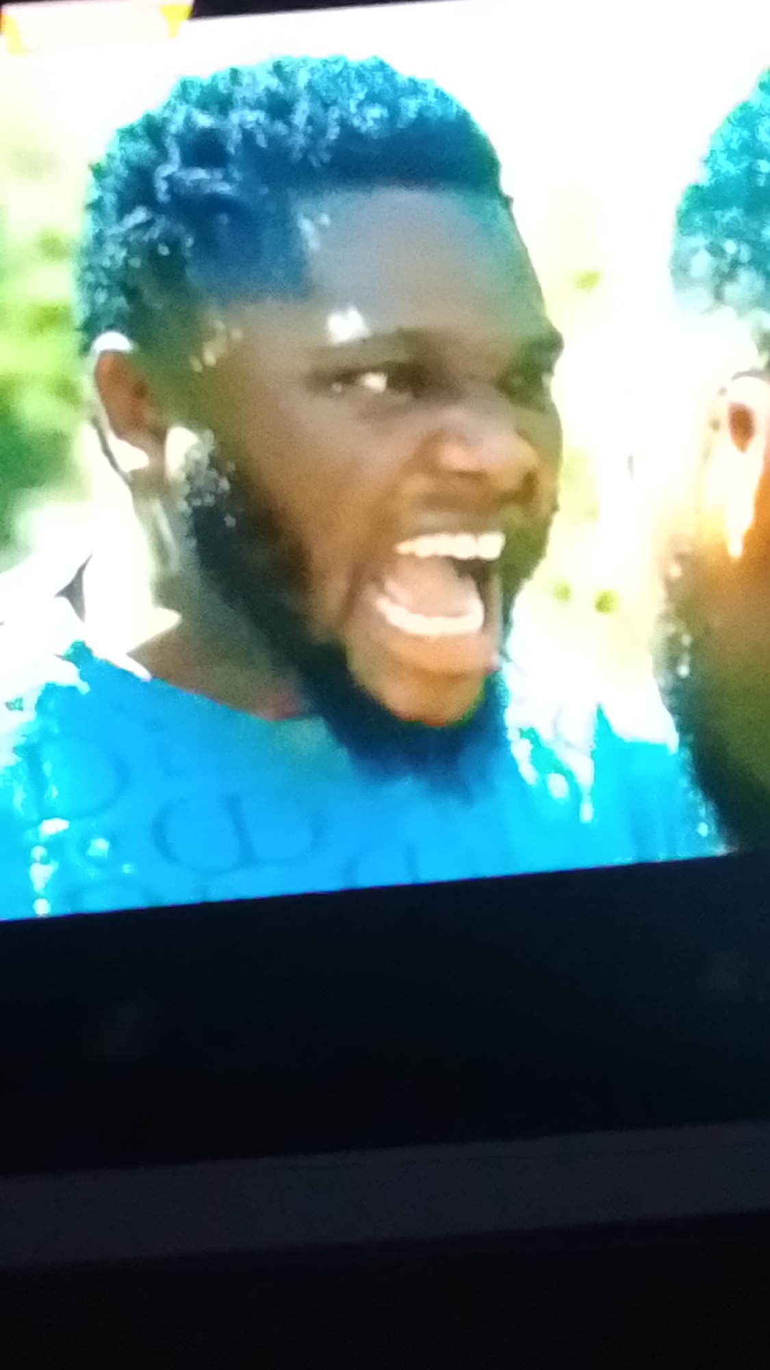 The image captures a close-up of a dark-skinned individual, likely male, from the chest up, positioned slightly off-center to the left. The person has short, tightly curled hair dyed a vibrant blue-green (teal) color and a dark beard. The individual's mouth is wide open, exposing upper and lower teeth, in what appears to be a scream or shout. Their eyes are open and directed towards the right side of the frame. The person is wearing a bright blue short-sleeved t-shirt, upon which subtle, embossed letters, possibly "CD" or similar, are visible. The background is an out-of-focus outdoor scene characterized by bright, natural light and blurred green foliage, indicative of a daytime setting. A portion of another person with similarly dyed blue-green hair is partially visible on the right edge of the frame. The specific location in Lokoja, Nigeria, cannot be determined from the visual evidence provided.