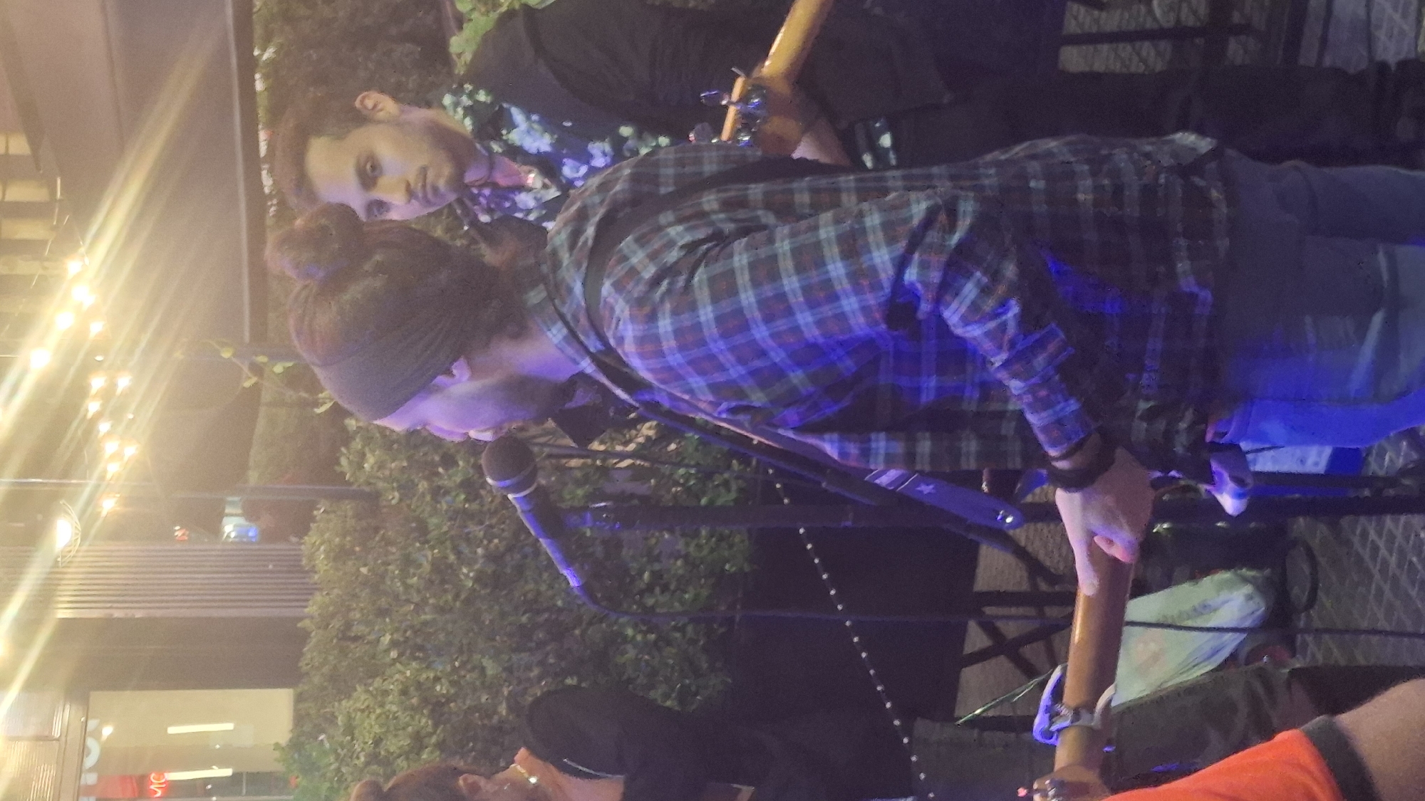A band is performing at night. One man with a black headband and wearing a plaid shirt plays a guitar and sings into a microphone. Another man stands behind him wearing a shirt with a floral pattern, and plays the guitar. Both are standing on a stage. Other patrons are visible. String lights are in the background. The location may be an outdoor venue in Buenos Aires, Argentina.