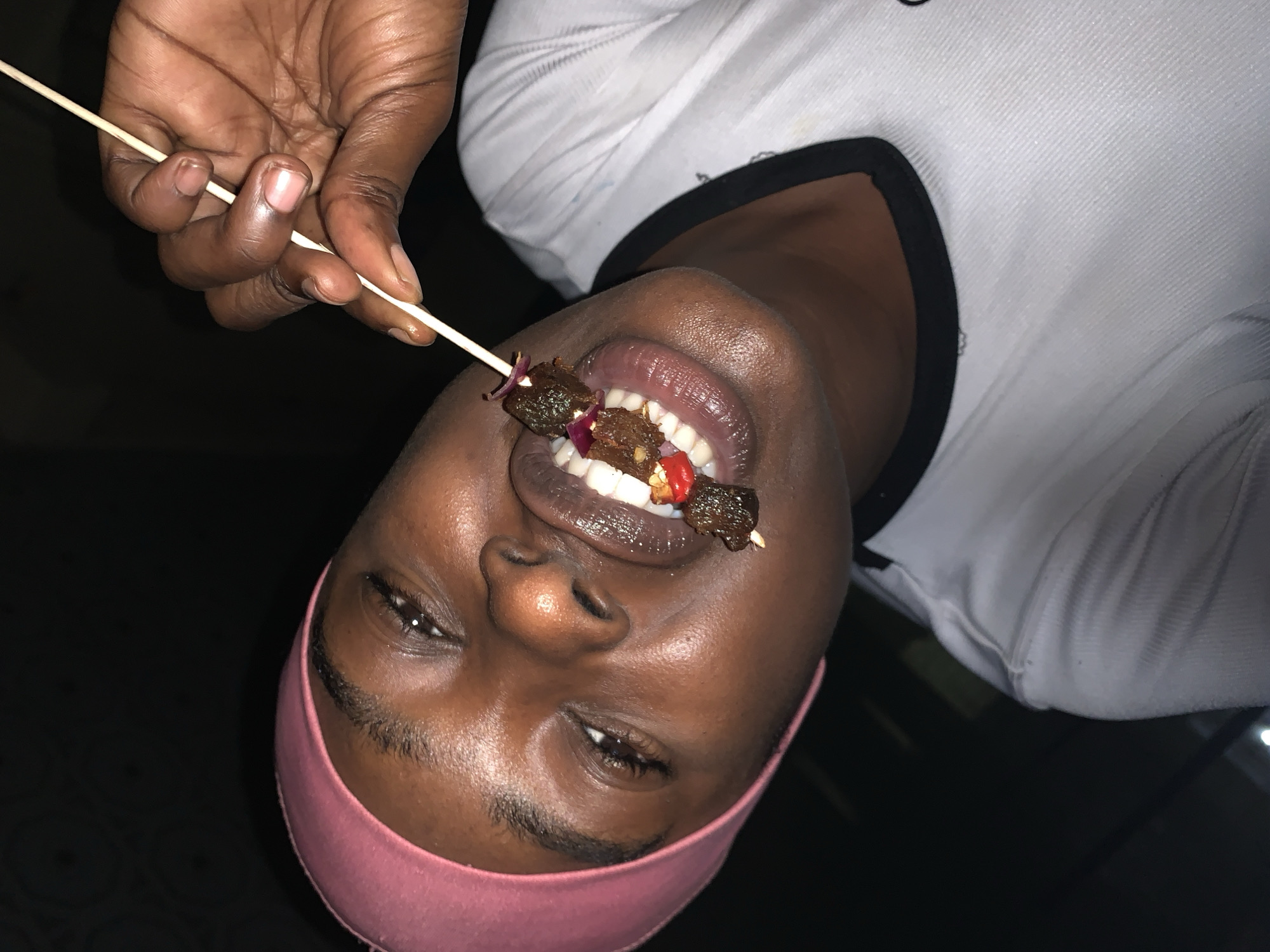 A person with dark skin and a pink headband is holding a skewer of food in their mouth. The skewer contains pieces of meat, red pepper, and onion. The person is wearing a white shirt with a black collar. The location is implied to be in the city of New Stadium, Nigeria. The person appears to be posing for a photo while eating.