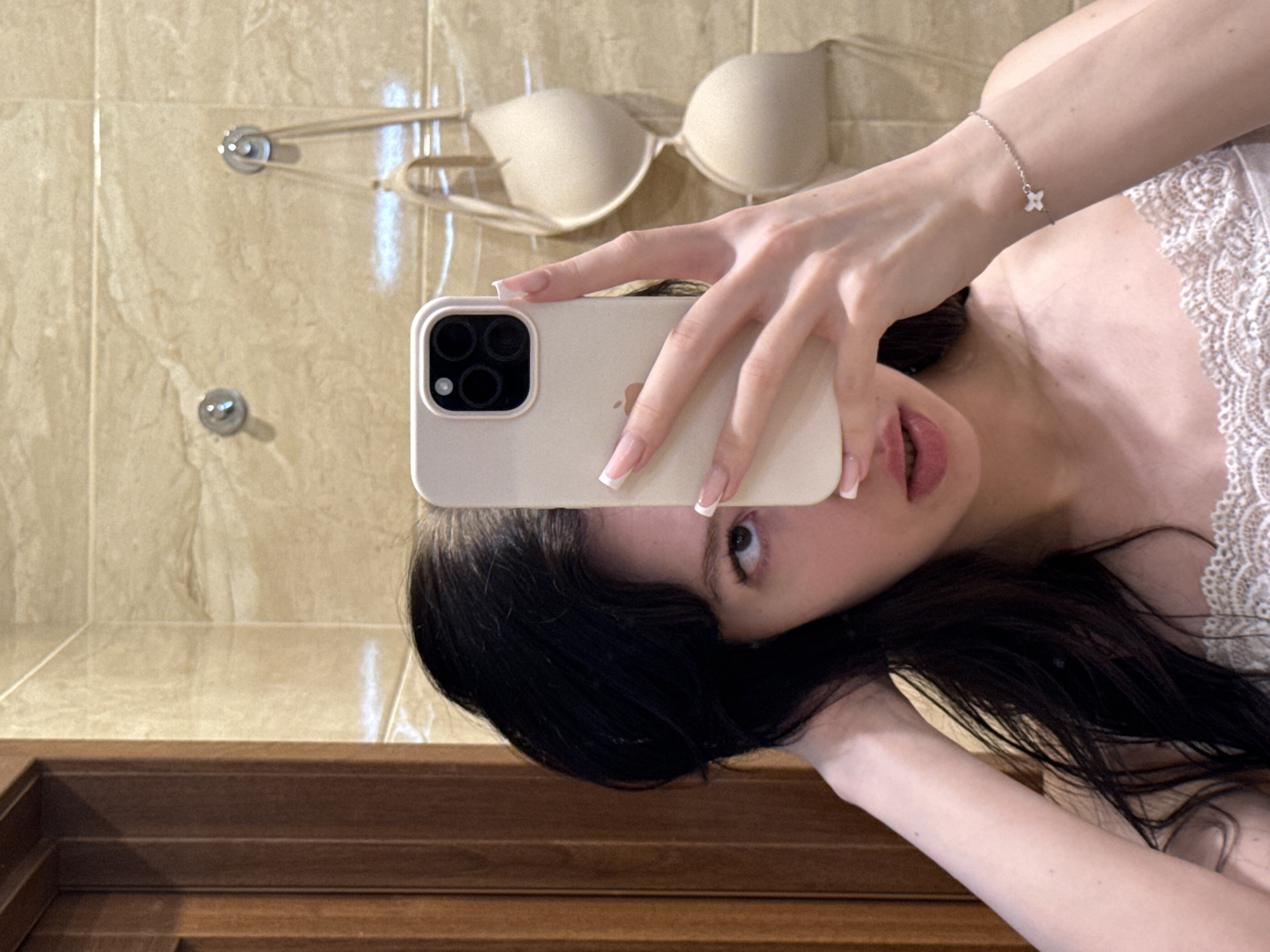 A young woman with dark hair is taking a mirror selfie in a bathroom. She is holding a light-colored smartphone in her right hand, covering the lower half of her face. Her left hand is touching her hair, which is falling around her shoulders. She is wearing a white lace-trimmed top. In the background, a beige bra is hanging on a hook on a tiled wall. To the left of the frame, a wooden door is visible. The scene appears to be indoors, likely in a private residence. The location of Khimki, Russia, is not explicitly indicated by any visual cues in the image.