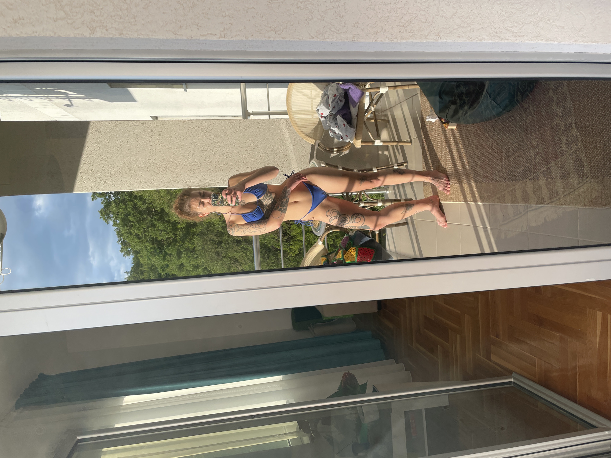 A woman is reflected in a window, taking a selfie on a balcony. She is wearing a blue bikini and has numerous tattoos on her arms and legs. The balcony has a table and chairs, a rug, and a view of green trees and a blue sky. The room inside the window has a light blue curtain and a herringbone wood floor. It is plausible that the location is Budva, Montenegro, due to the architectural style and coastal environment, but this cannot be confirmed from the image alone.