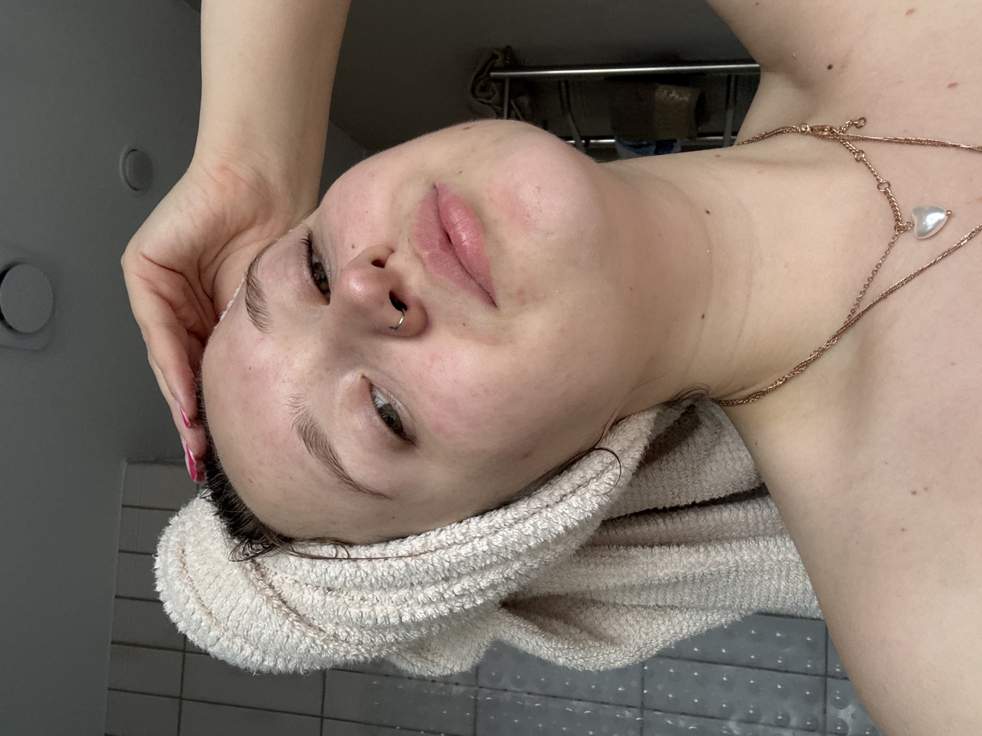 A young person, possibly female, with fair skin, is lying on a tiled surface, likely a bathroom floor. The person's head rests on a white terry cloth towel. A gold chain necklace with a pearlescent pendant is visible on the neck. The person has a nose ring in their left nostril and their right hand is touching their forehead. The background includes a bathroom wall with fixtures such as a towel rack. The location cannot be determined as Izhevsk, Russia, from the image.