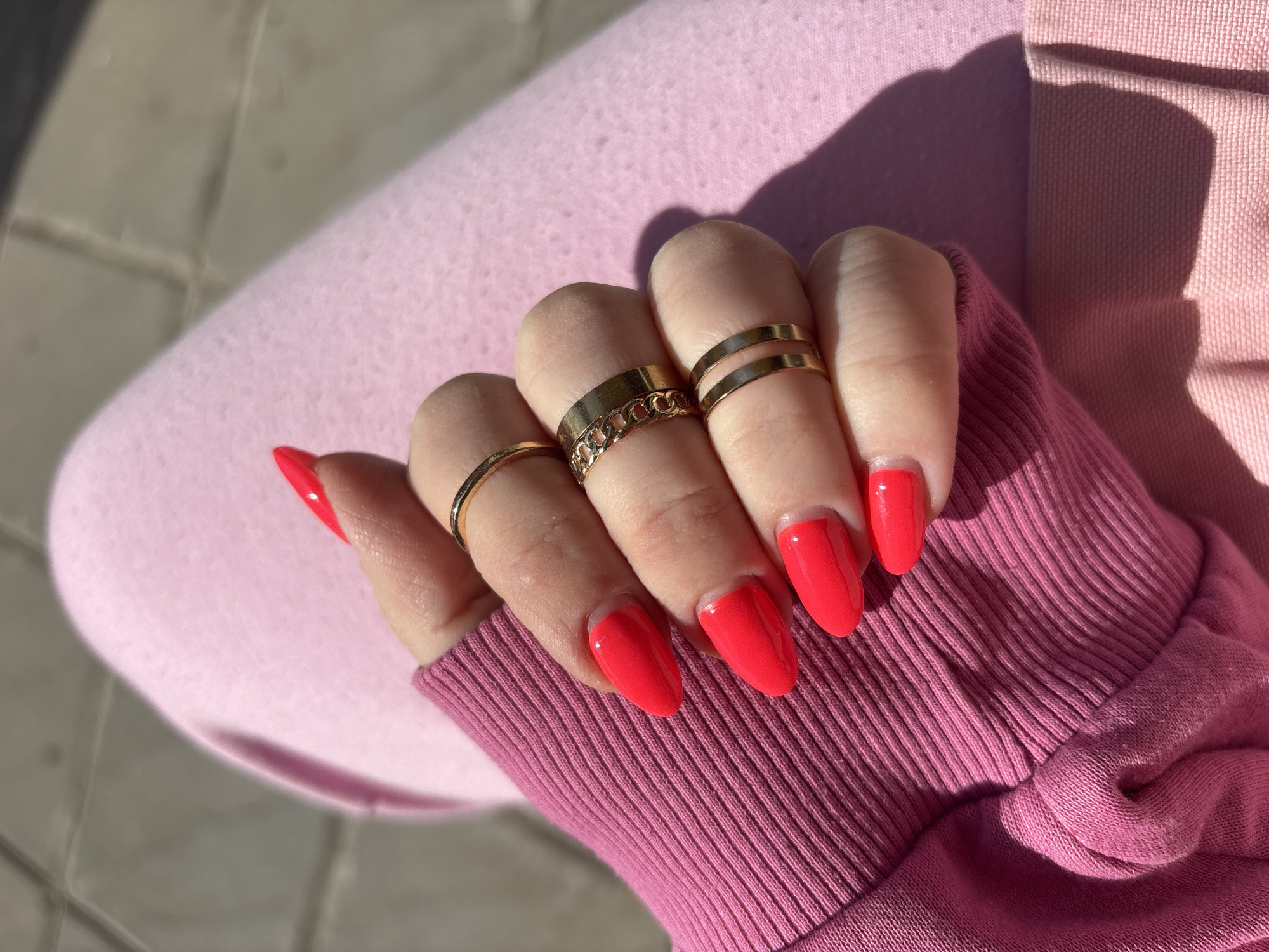 A close-up shot focuses on a person's left hand, adorned with vibrant red, glossy nail polish and gold-toned rings. The hand is positioned over textured pink fabric, likely the sleeve of a sweatshirt or hoodie, with a ribbed cuff prominent in the foreground. Another area of lighter pink fabric is visible beneath the hand, possibly part of another garment.
The nails are almond-shaped and painted a vivid, almost neon, red color. The hand wears four rings: a simple gold band on the pinky finger, a gold ring with a chain-link design on the ring finger, and two stacked plain gold bands on the middle finger.
The scene is set outdoors, under bright, direct sunlight that casts distinct, sharp shadows across the pink fabric, including a shadow from the hand and arm. The background reveals a light gray concrete or paved surface with visible lines, suggesting an urban or outdoor pathway. The strong light indicates it is daytime, likely morning or late afternoon. The overall impression is casual and stylish, captured in a sunny outdoor setting in Sofia, Bulgaria. No text is visible in the image.
