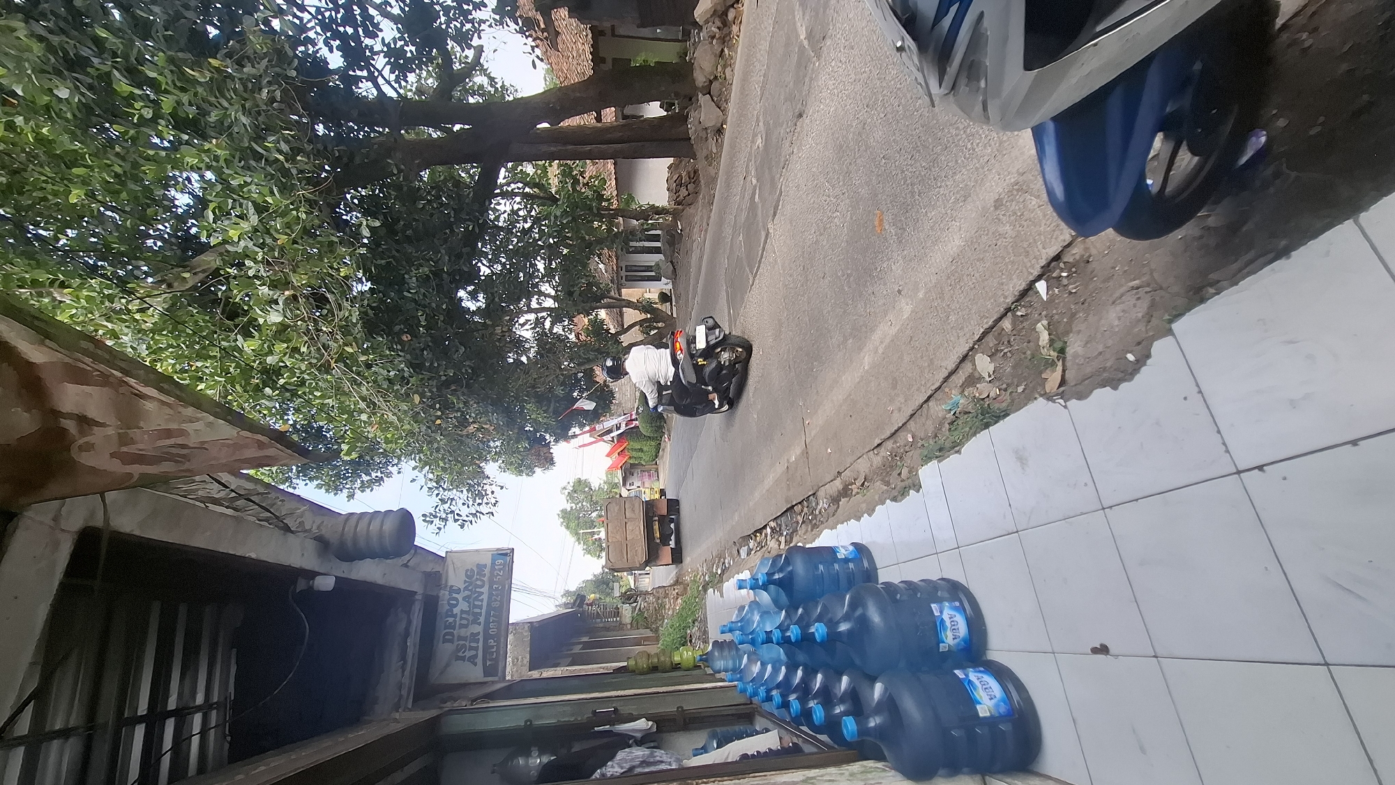 A street scene in Cilaku, Indonesia, features a row of large blue water jugs lined up on a tiled sidewalk in front of a building with a corrugated metal roof. A sign on the building reads "DEPOT ISI ULANG AIR MINUM" with a phone number below. A dump truck is visible on the paved road in the background, followed by a person riding a black scooter. The road is lined with trees and houses on either side. A portion of a motorcycle is in the foreground, on the right side of the image.