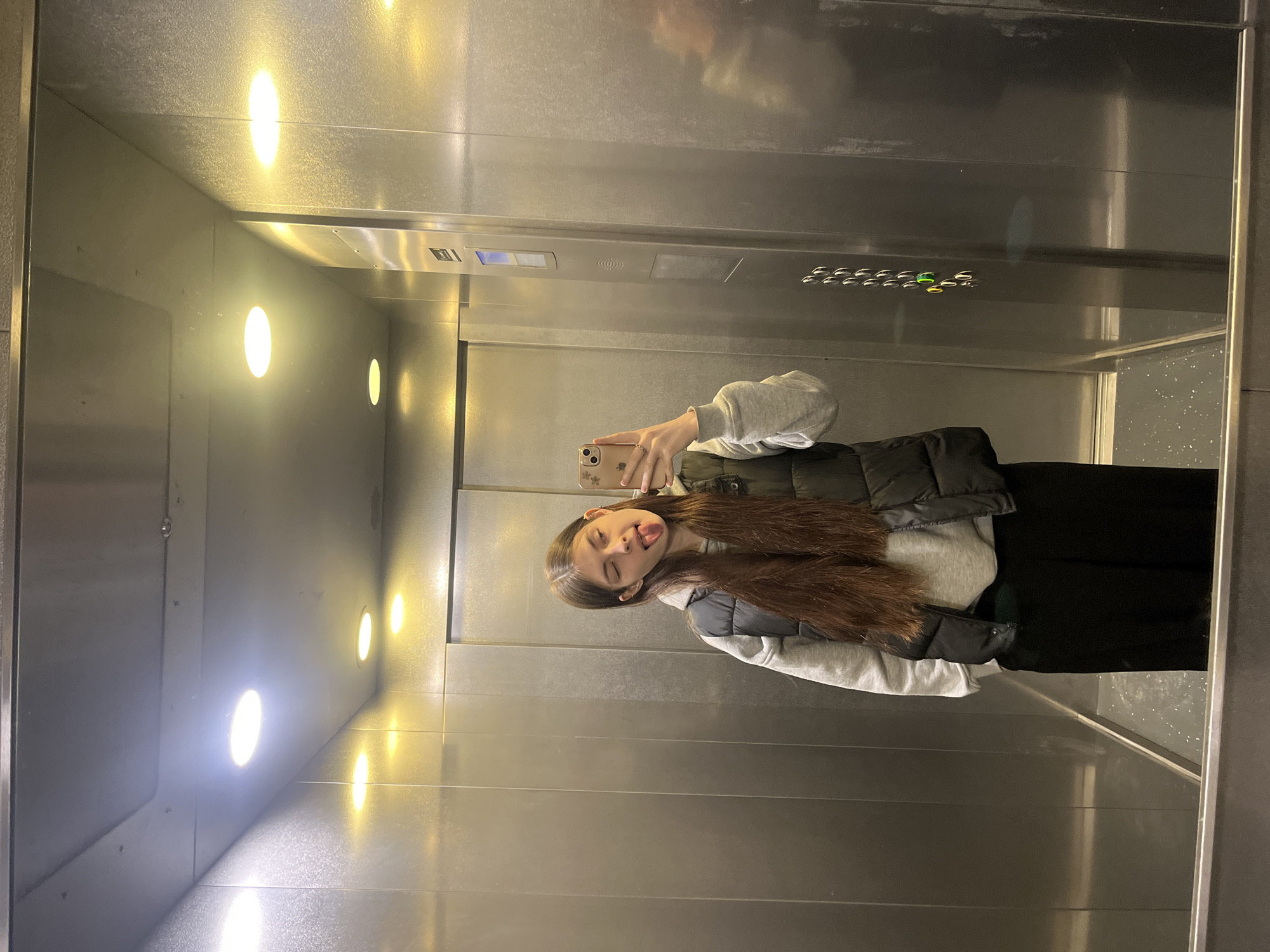 A young woman is taking a selfie in an elevator. She is wearing a grey sweatshirt, a dark vest, and black pants. She has long brown hair. She is sticking her tongue out at the camera. The elevator has shiny metal walls with several round lights. The control panel can be seen above her. The elevator's mirror surface reflects her image. The location appears to be Saint Petersburg, Russia, based on the modern elevator design common in European countries, including Russia.
