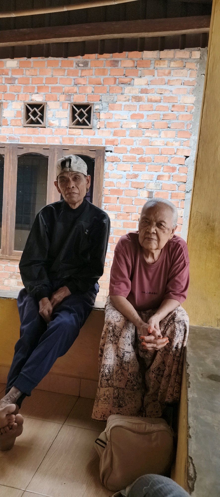 In Muaro Jambi, Indonesia, an elderly Indonesian couple sits on a raised ledge outdoors, likely a veranda or porch. The man on the left wears a black jacket and dark blue pants, with a light-colored cap on his head. The woman on the right is dressed in a maroon t-shirt and a patterned skirt. Both individuals have their hands resting in their laps. In front of them, on the tiled floor, is a light-brown bag. The background features a brick wall with two square ventilation grates, and a wooden-framed window. The overall setting appears to be residential, given the humble construction and the presence of a couple in a relaxed, outdoor setting.