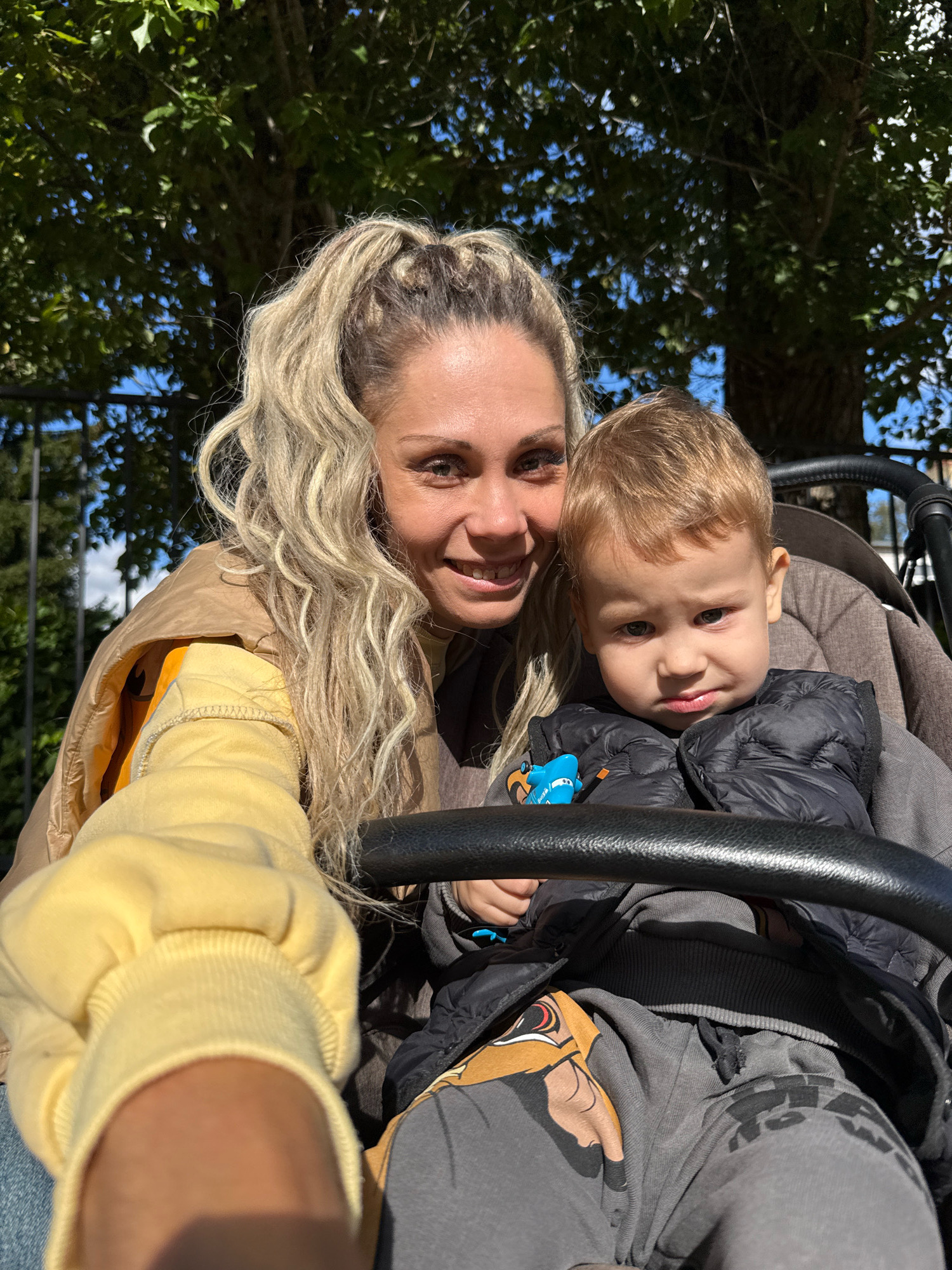 In Rusanovo, Russia, a woman with blonde hair pulled into a ponytail is taking a selfie in an outdoor setting, possibly a park, as indicated by the visible trees and dappled sunlight. She is wearing a yellow hoodie and a tan jacket over it. Next to her, in what appears to be a stroller, is a young child with blonde hair. The child is wearing a black puffer vest over a grey hoodie. The child is holding a small blue toy. The focus is on the woman and child, with the background blurred, suggesting a shallow depth of field. The overall mood appears casual and familial.