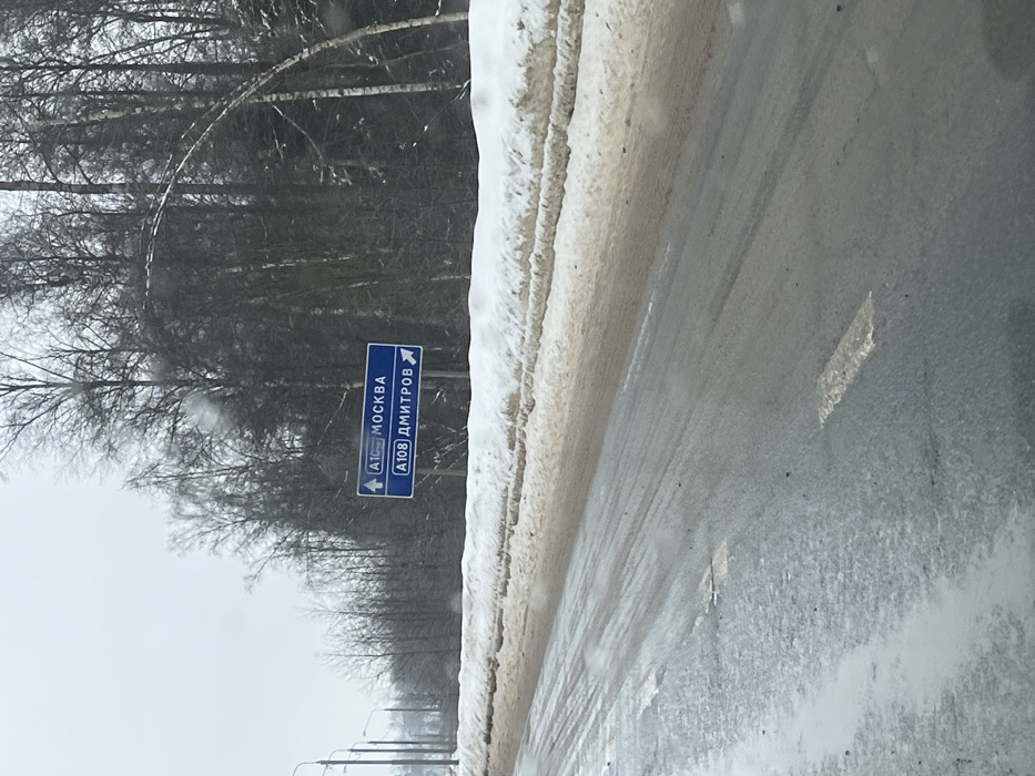 The image depicts a winter road scene from the perspective of a vehicle, looking forward. The road surface is dark, wet, and appears slushy or icy in patches, showing visible tire tracks and faint, worn white lane markings. Dirty, brown-tinged snow banks line both sides of the road, with a more substantial accumulation on the right. Beyond the snow banks, a dense line of bare deciduous trees, primarily birches, extends into the background, indicating a forested area. The sky is overcast and uniformly light gray.
On the right side of the road, a blue rectangular road sign is mounted on a pole. The sign contains two white panels:
1. The upper panel displays "A108 МОСКВА" (A108 MOSCOW) with an upward-pointing arrow, indicating that continuing straight on the A108 leads towards Moscow.
2. The lower panel displays "A108 ДМИТРОВ" (A108 DMITROV) with a rightward-pointing arrow, indicating that a right turn leads onto the A108 towards Dmitrov.
Utility poles with streetlights are visible receding into the distance along the left side of the road. No individuals or active interactions are visible in the scene.
The location is on the A108 (Moscow Big Ring Road), specifically at a junction or fork in the road near the village of Ivashevo, located in the Dmitrovsky District of Moscow Oblast, Russia. The sign directs traffic on the A108, with one path designated for Moscow (straight ahead) and another for Dmitrov (right turn).