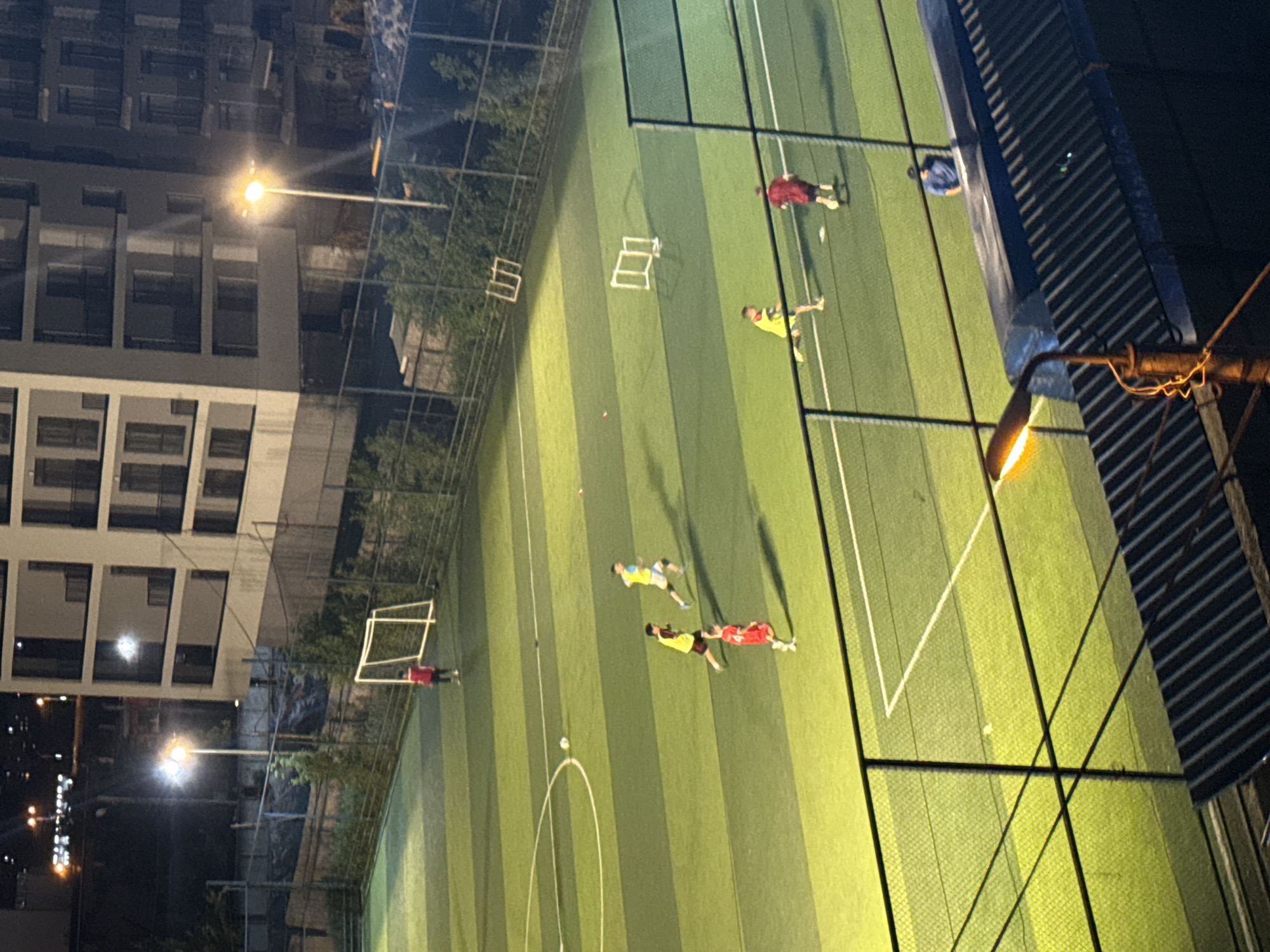 This is a nighttime aerial photograph of a soccer field in Tbilisi, Georgia. The field is illuminated by bright lights, casting long shadows. There are several people playing soccer on the field. One person is standing near a small goal on the left side of the image, wearing a red shirt. On the field, four other individuals are visible in various poses related to playing soccer. Two are wearing yellow shirts and running, one is in a red shirt and is also running, and another is in a dark shirt, appearing to be kicking a ball. The field is marked with white lines and has a synthetic turf surface. Behind the field, a multi-story building with balconies is visible, along with streetlights and trees. A fence separates the field from the viewer's vantage point, with a dark, corrugated roof structure in the foreground.
