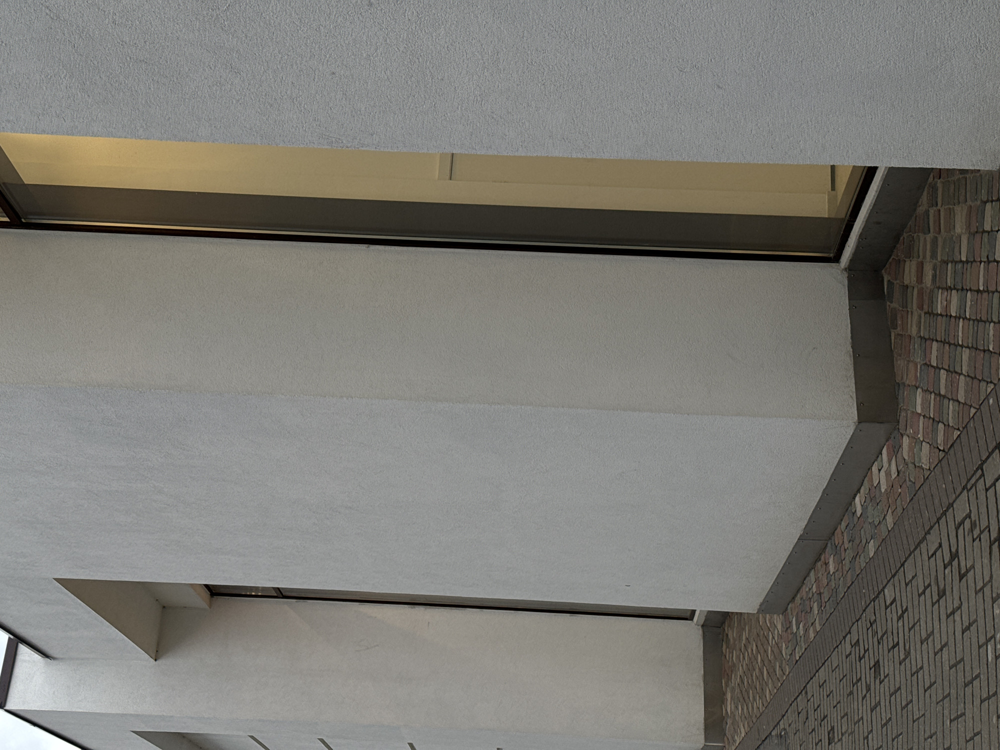 The image captures an upward-looking view of modern building architecture, dominated by light gray concrete structures, a window band, and a textured brick wall.
Large, textured light gray concrete slabs form a ceiling at the top and a prominent horizontal overhang in the mid-ground. These concrete surfaces exhibit a slightly coarse, granular finish. Between the upper ceiling and the lower overhang, a long, narrow horizontal window or opening is visible. Through this window, a warm, yellowish light is reflected or visible from an interior space, suggesting illumination from within. The window frame is dark, likely metal.
On the right side of the image, a multi-colored brick wall extends upwards. The bricks display varying shades of brown, gray, and some reddish tones, arranged in a mix of patterns, including a section resembling a basket weave or herringbone pattern at a lower level, transitioning to a more standard bond above, separated by a darker horizontal brick band. A dark metal trim piece connects the concrete overhang to the brick wall.
The lighting appears diffuse, possibly indicating an overcast day or the scene being in shade, contrasting with the bright light seen through the windows. There is no discernible activity, people, or visible text in the image. The scene is static, focusing on the structural and material details of the building exterior.